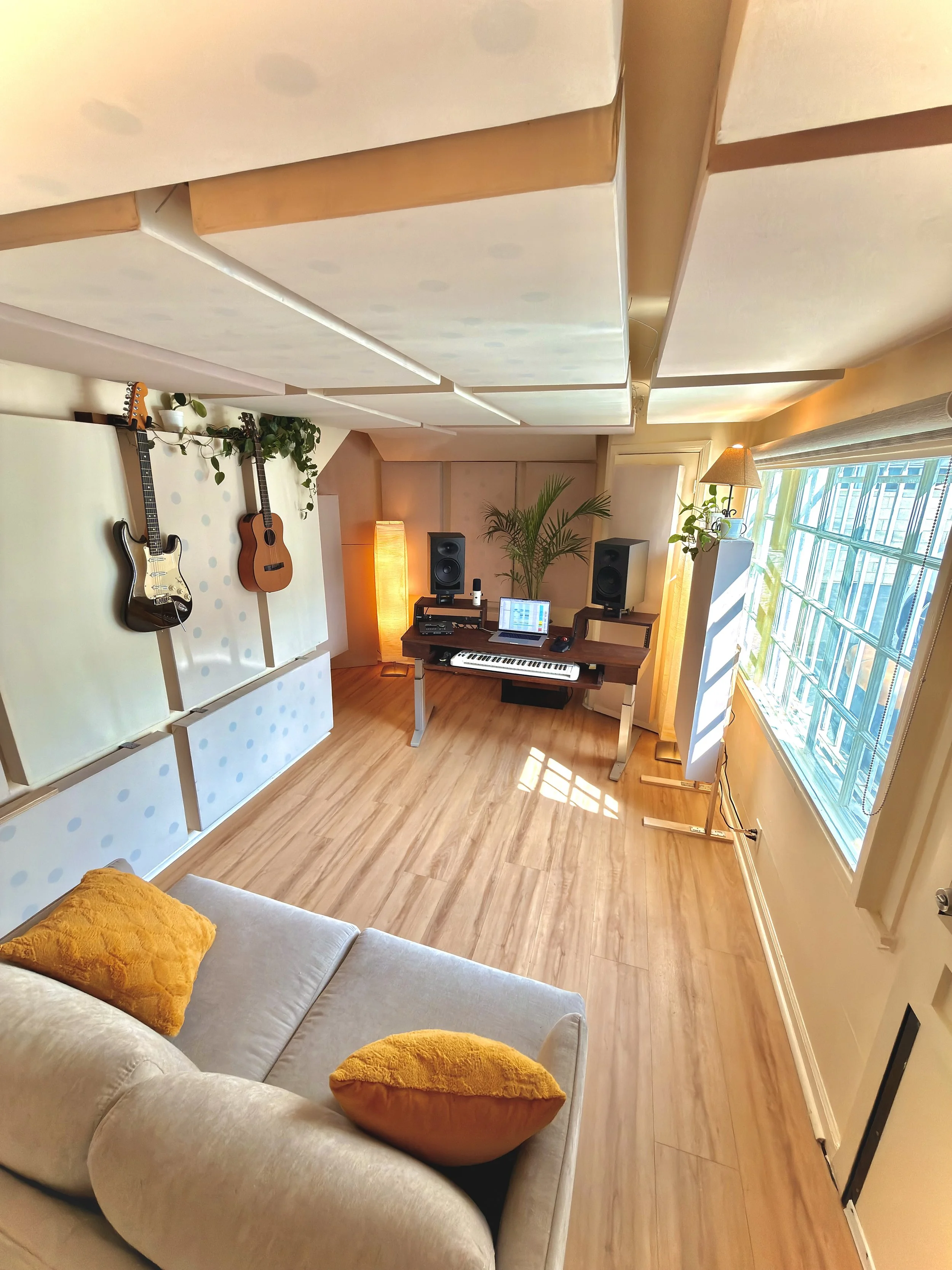 View of a cozy indoor music studio with a white couch and orange pillows, a wall-mounted music stand with guitars, a keyboard, laptop, speakers, and a potted plant near a large window with sunlight.