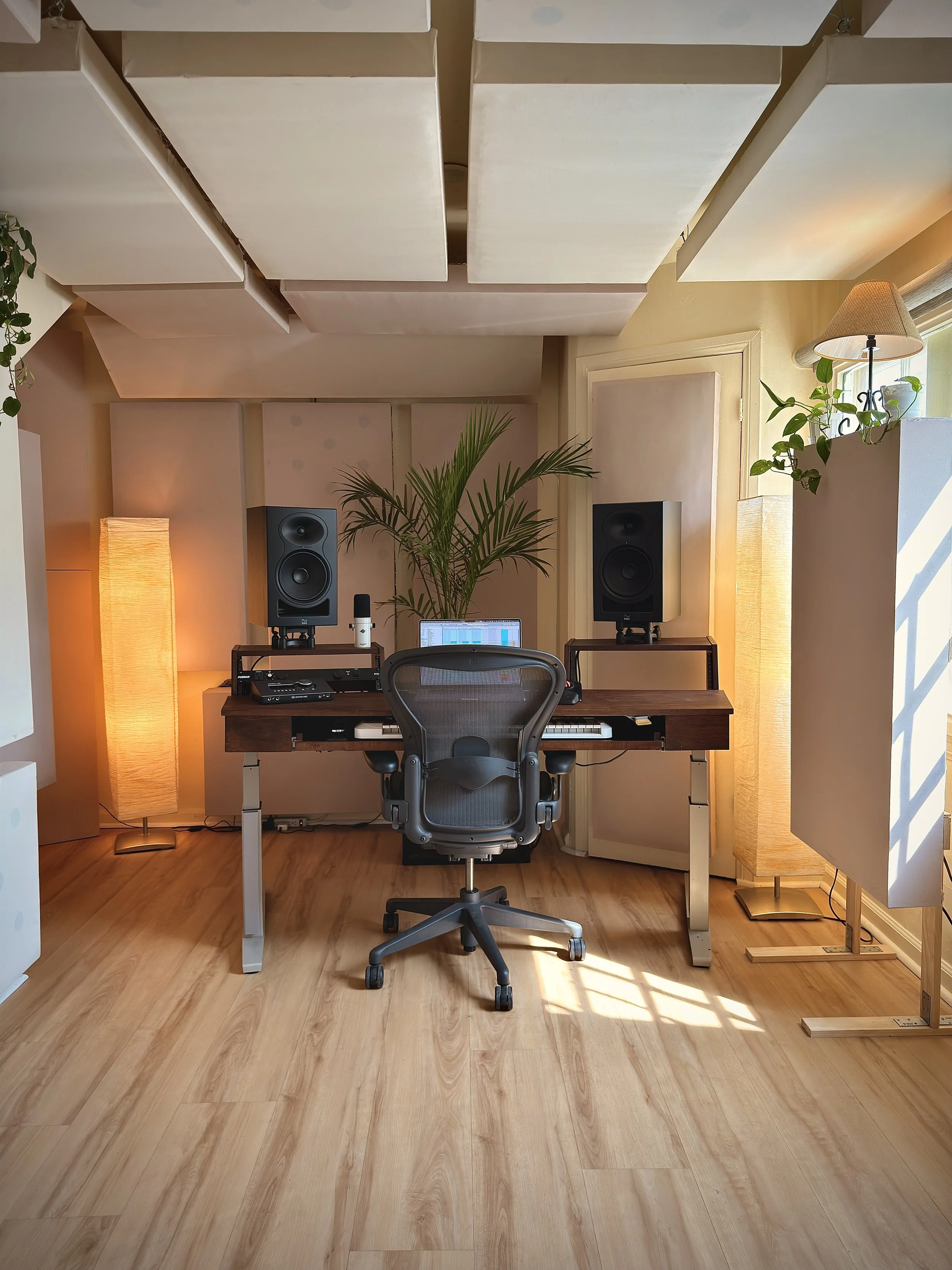 A recording studio with a desk holding audio equipment, two large studio monitors on stands, a computer screen, a white MIDI keyboard, and two tall floor lamps; black office chair in front of the desk, with plants near the window and acoustic panels on the ceiling.