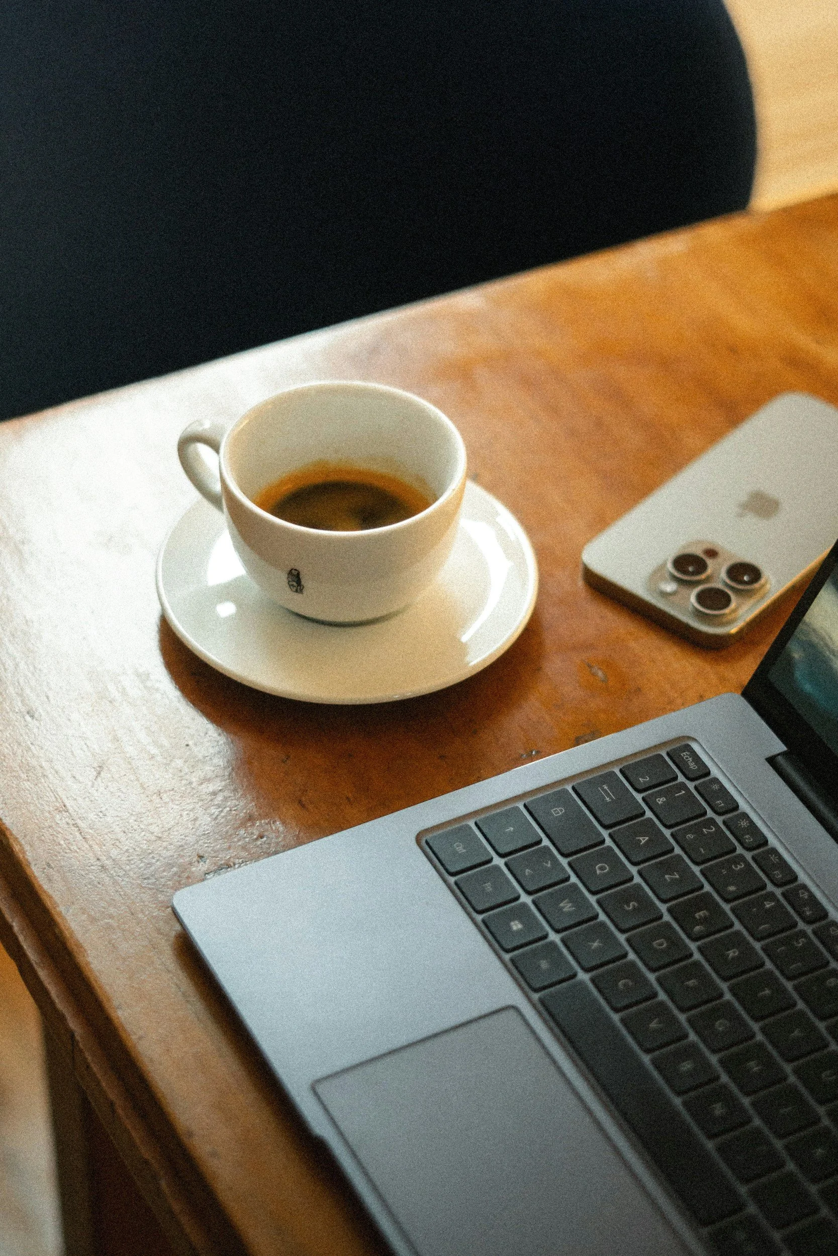 A wooden desk holds a silver laptop keyboard, a white cup of black coffee on a saucer, and an iPhone with a triple camera on the back. Interviewing guide for coffee shop owners and managers.