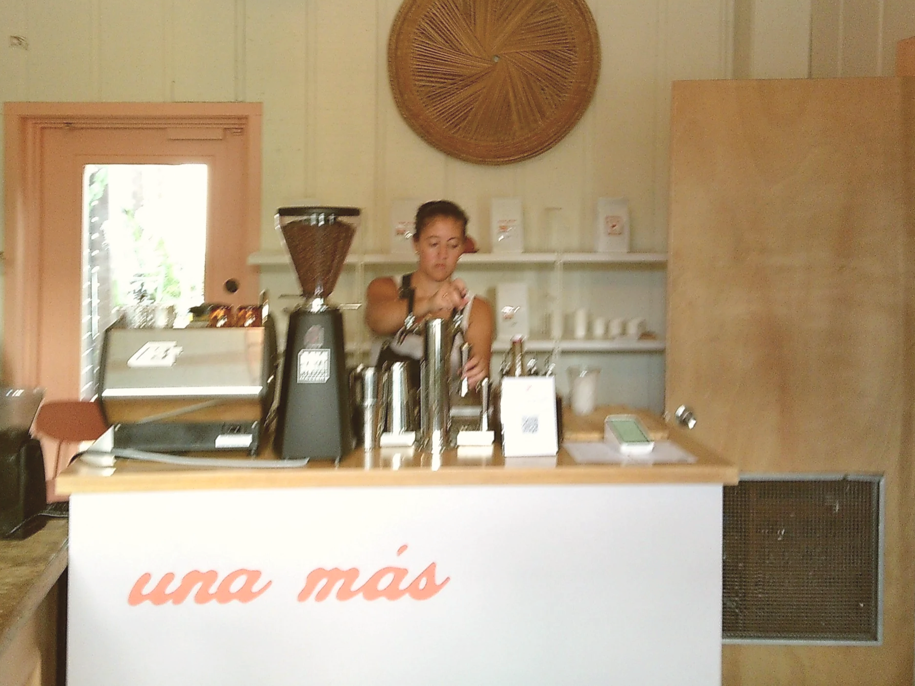 A woman behind a coffee cart preparing a drink, with coffee equipment, a cash register, and shelves with coffee products in the background. Paola Chamorro Ward. Una Más TPA