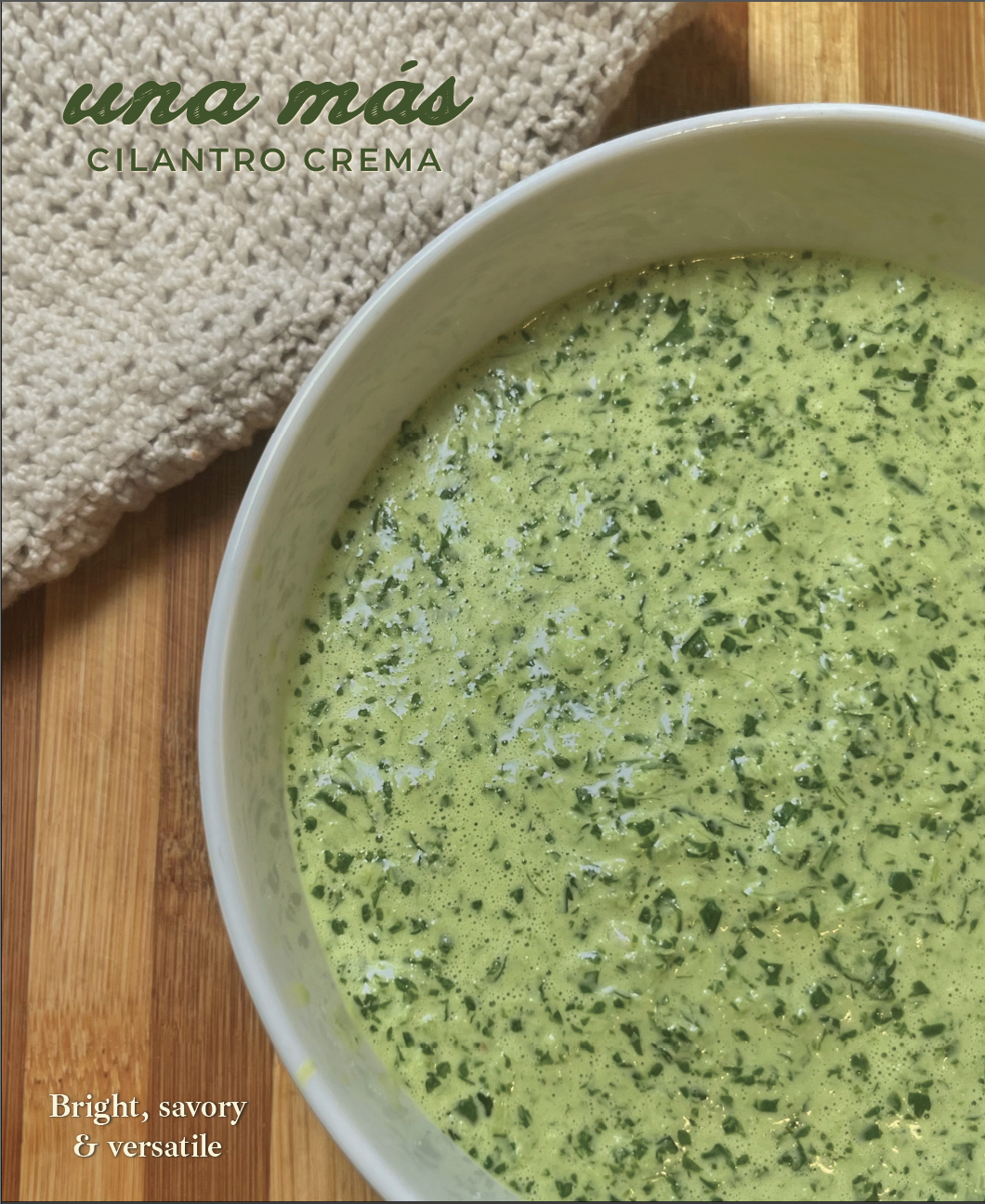 A bowl of cilantro creama sauce placed on a wooden surface. A beige knitted cloth is visible in the background. Text on the image reads 'una más' in green cursive and 'CILANTRO CREMA' in uppercase. Additional text says 'Bright, savory & versatile'.