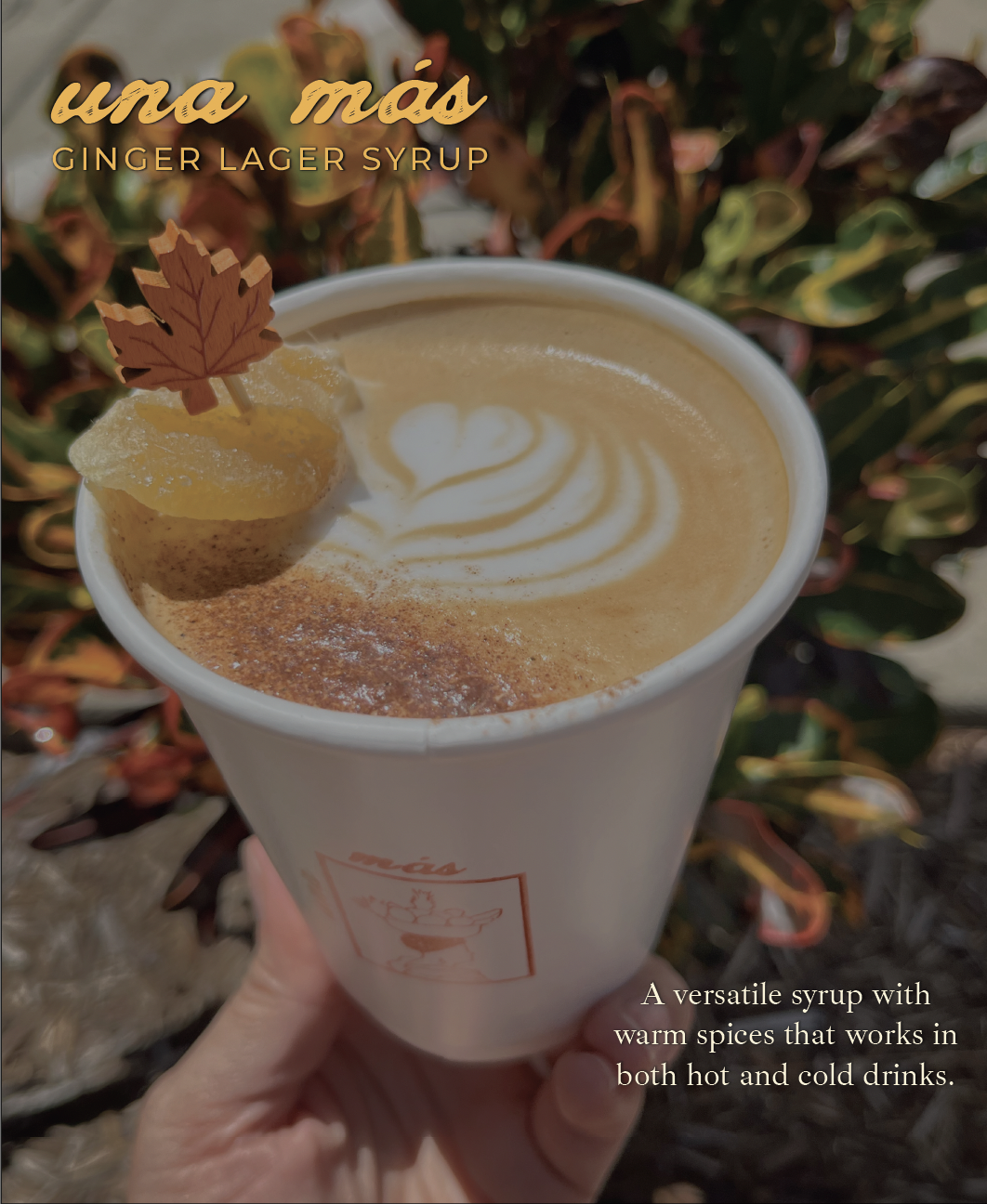 A hand holding a cup of a fall signature latte made with ginger lager syrup. It's outdoors with autumn leaves in the background, topped with latte art, a sprinkle of cinnamon, and a candied ginger on a leaf-shaped pick as garnish.