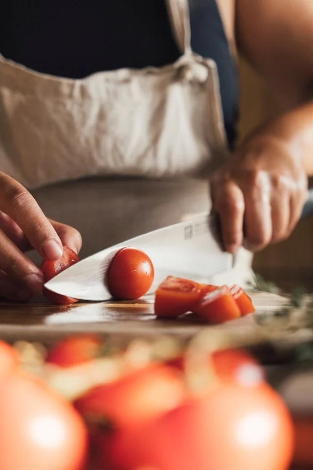 Person slicing a tomato on a cutting board with a chef's knife, with whole tomatoes in the foreground.