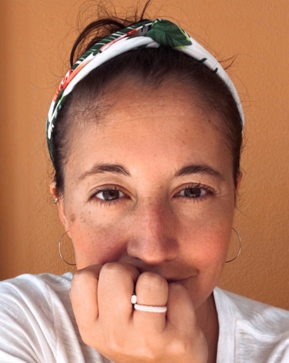 A woman with a bandana on her head and hoop earrings, smiling with her hand near her mouth, wearing rings, against a plain brown background.