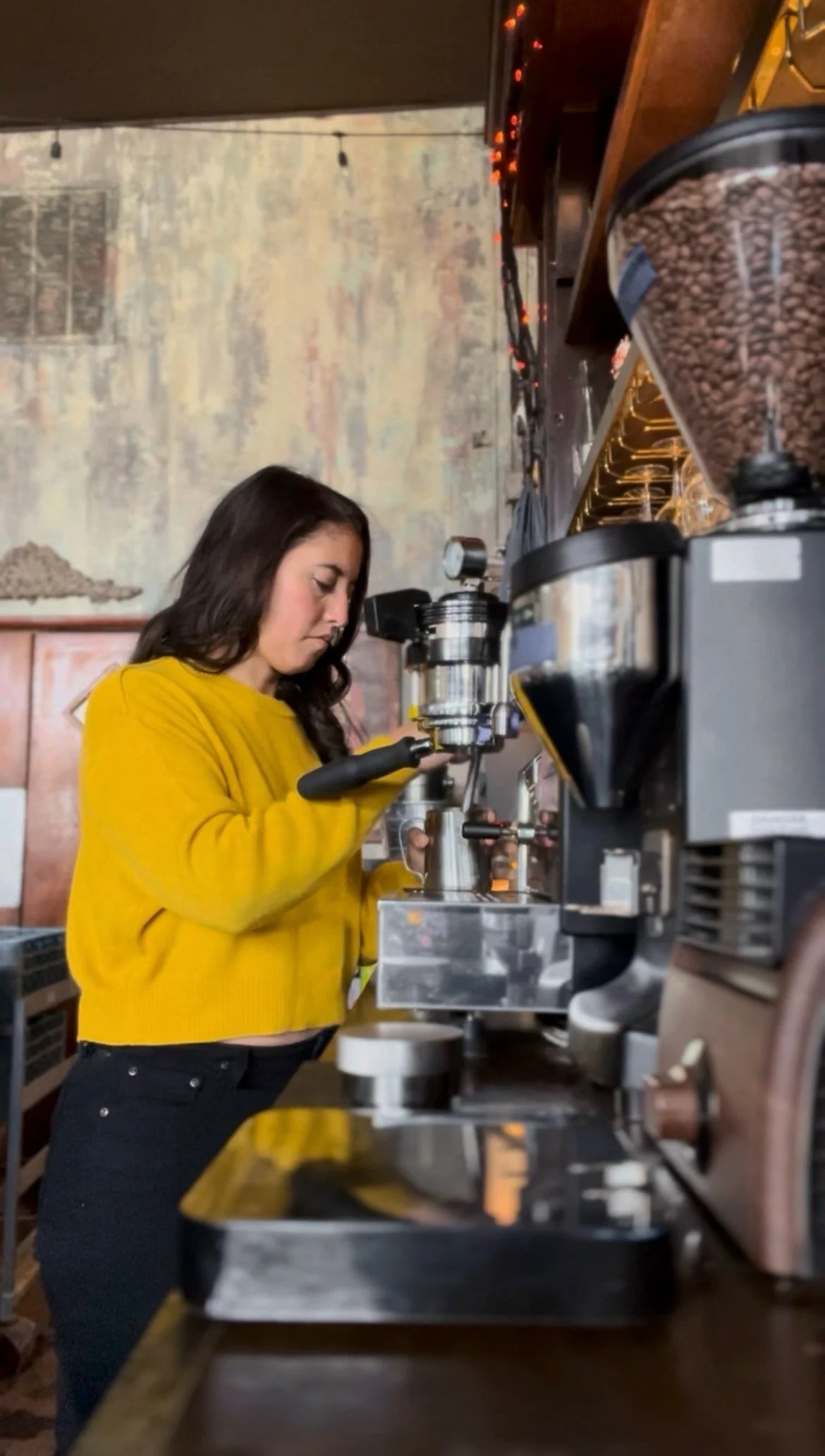 A woman in a yellow sweater making coffee using an espresso machine in a cafe.