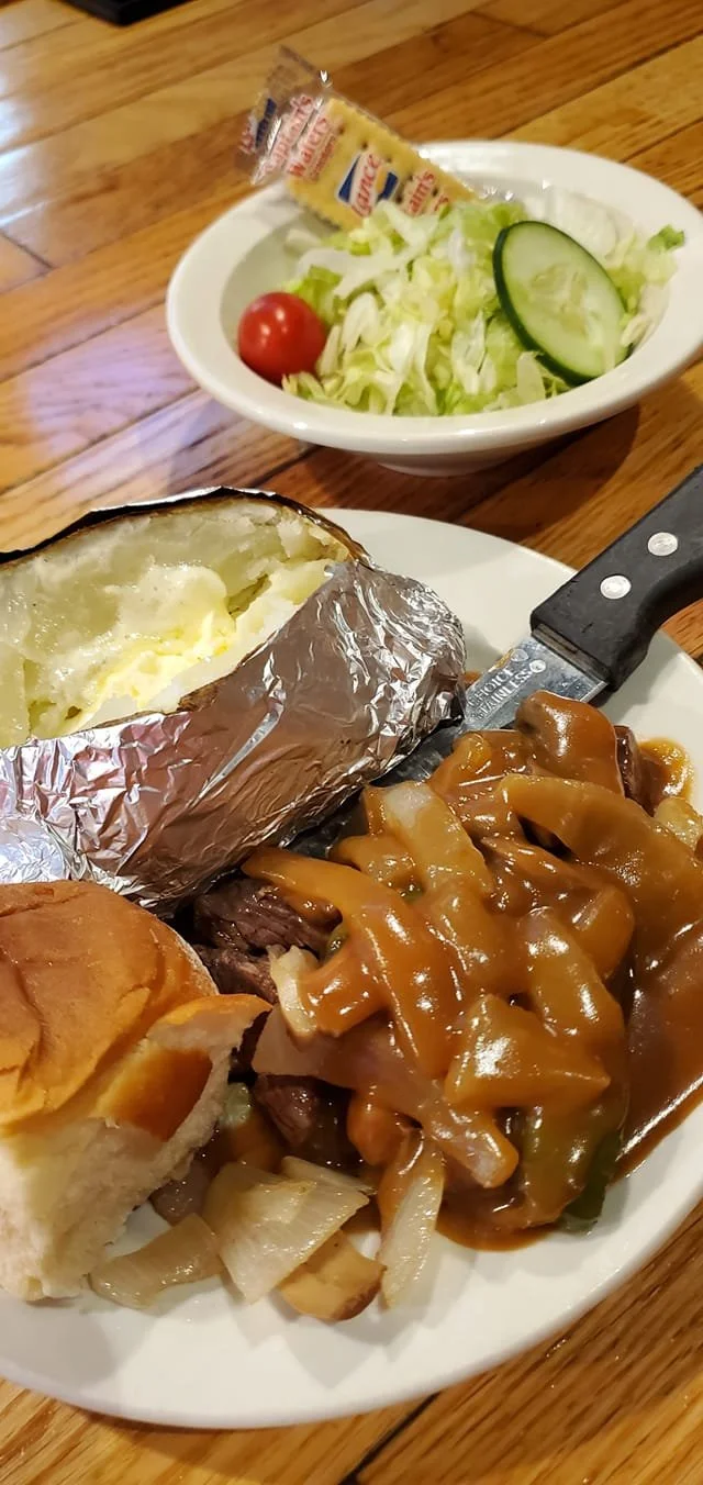Steak with gravy, baked potato with butter, side salad with lettuce, cucumber, cherry tomato, and a packet of salad dressing