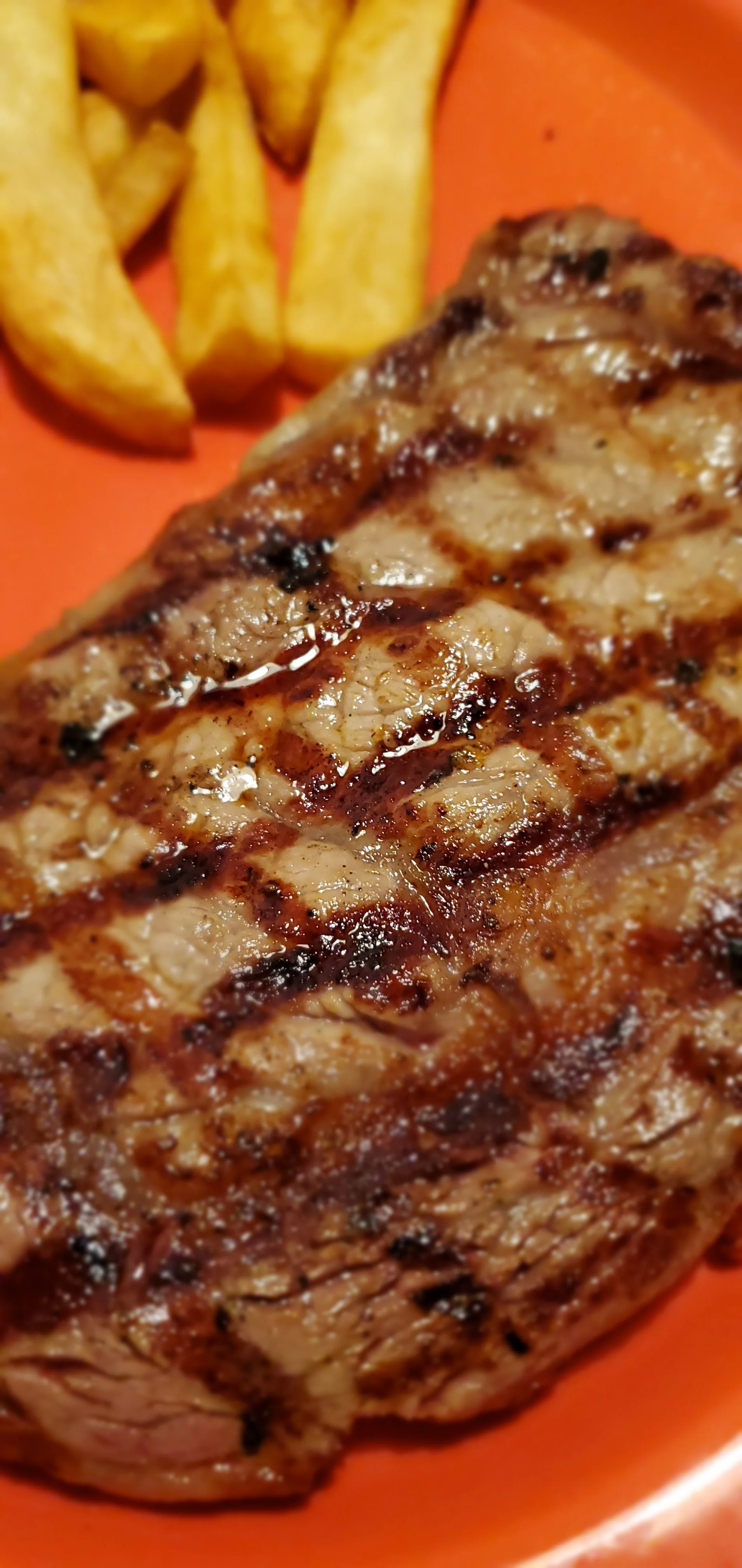 Close-up of a grilled steak with visible grill marks and a side of French fries on an orange plate.