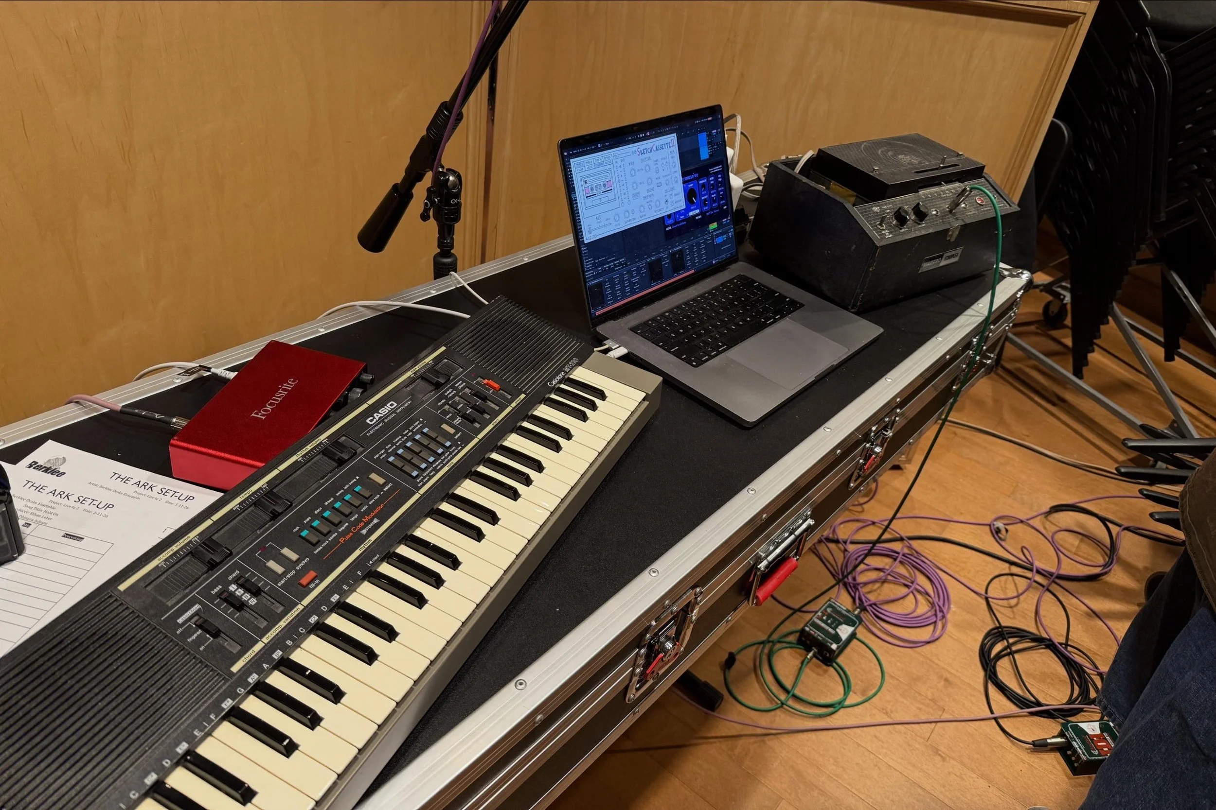 A music setup on a table including a keyboard, a laptop with digital music software, a small mixer, a microphone on a stand, and various cables and pedals in the background.