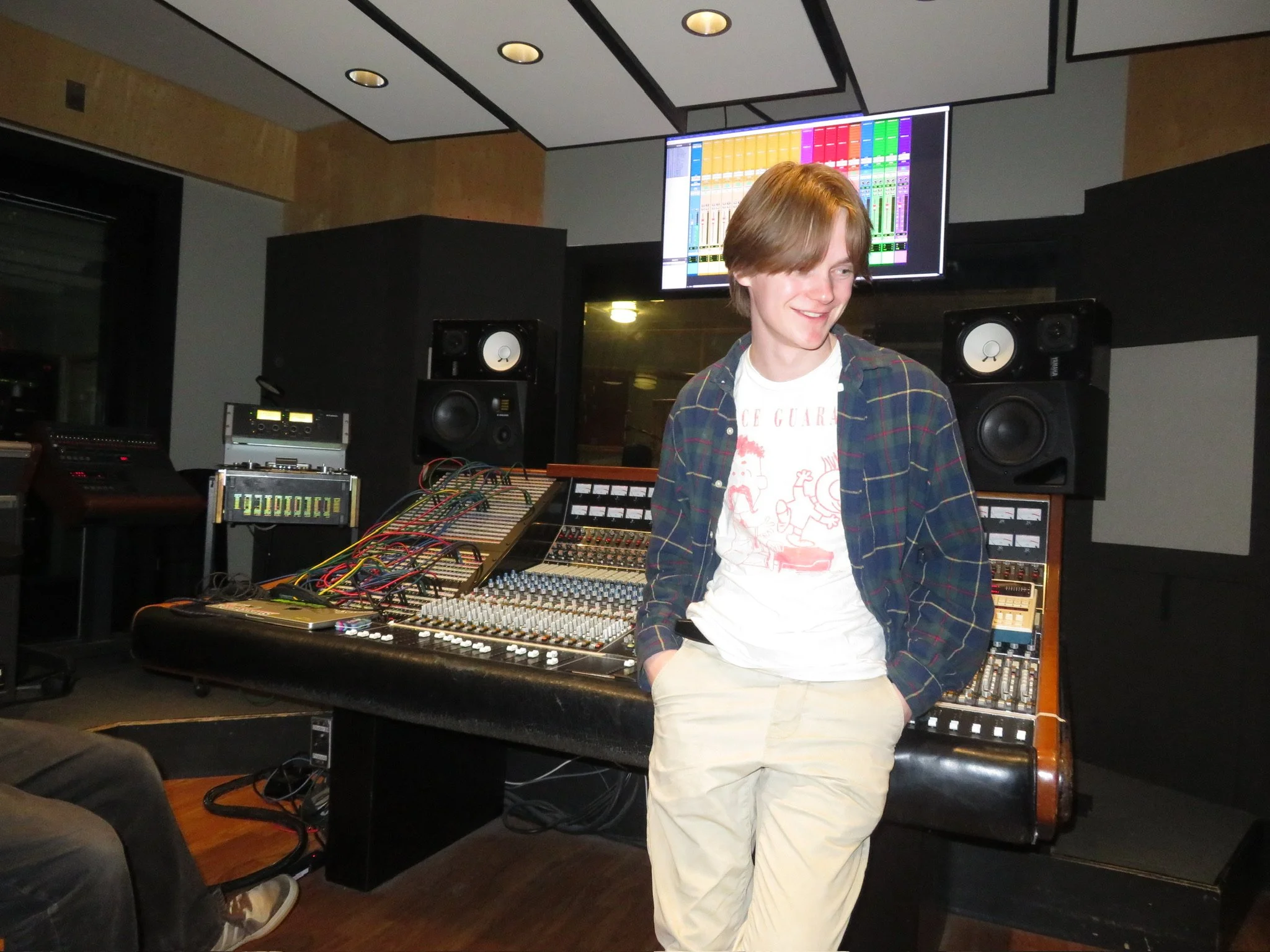 A young man with reddish hair smiling while standing in a music studio surrounded by professional audio equipment including large speakers, a mixing console, and a wide screen display showing digital audio workstations.