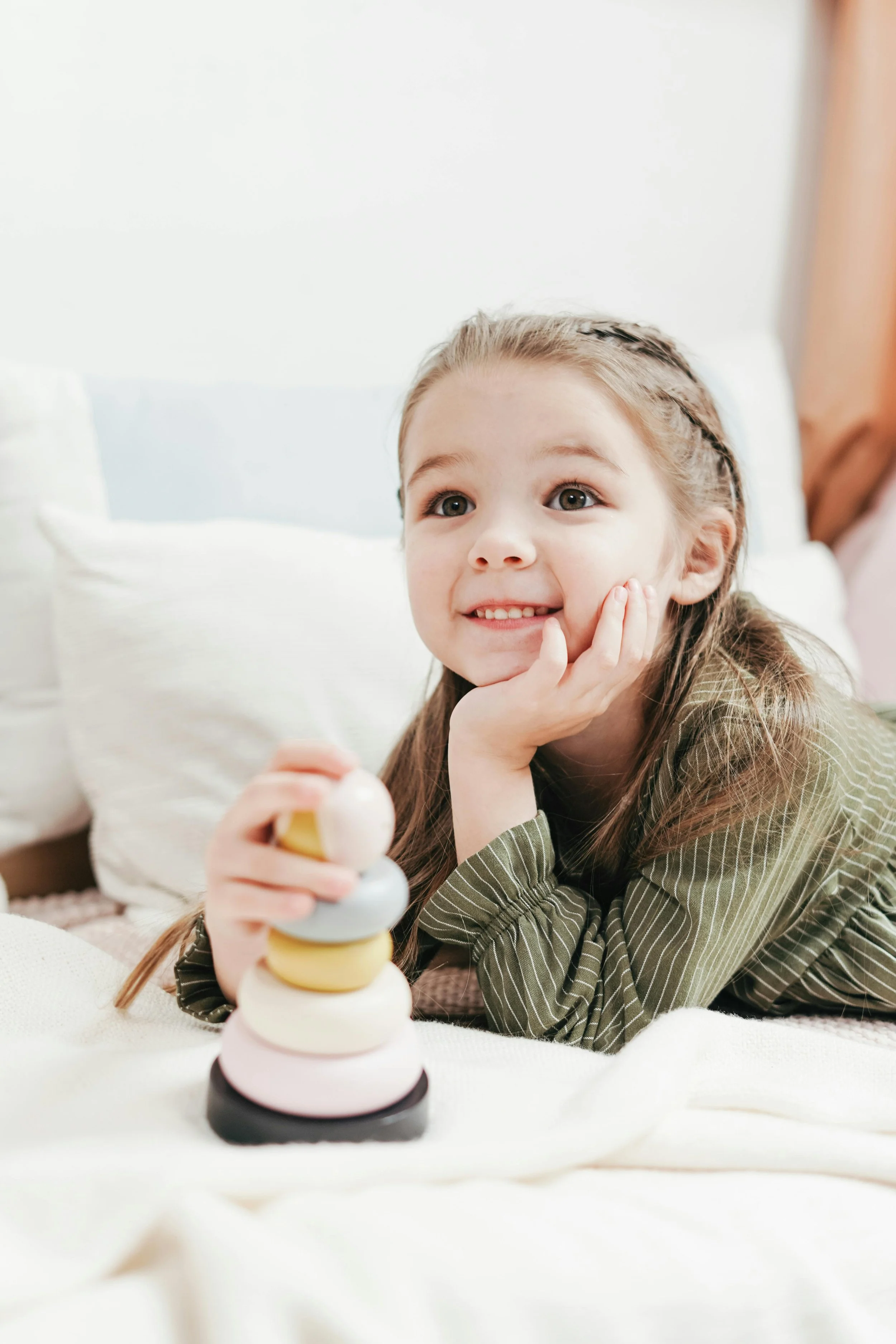 A young girl with brown hair and big eyes, smiling and resting her chin on her hand, playing with a stacking toy with colored rings on a bed or soft surface.