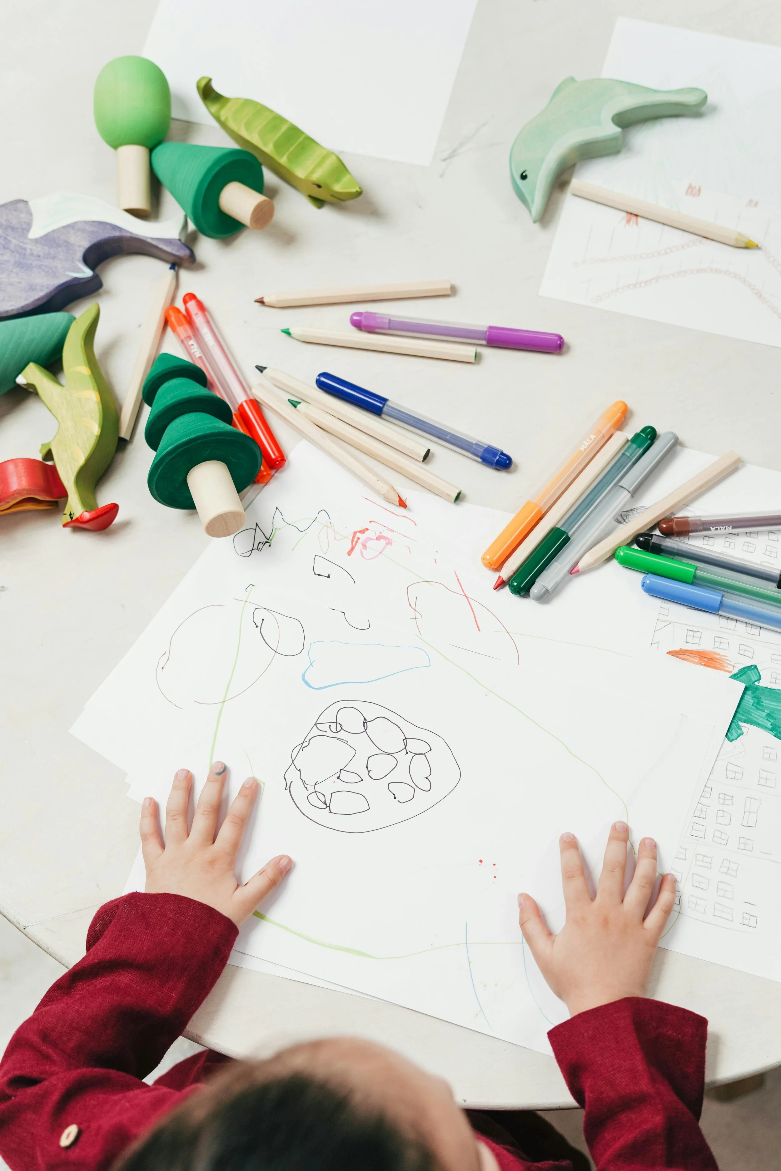 Children's hands on a table with colorful pens and drawings, including a cake sketch and abstract scribbles.
