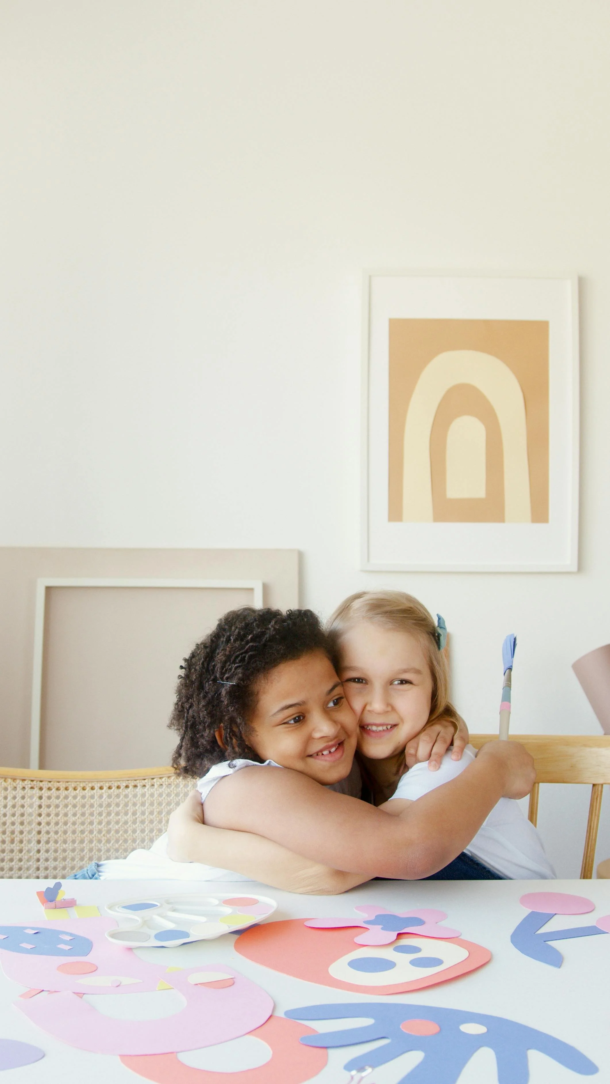 Two young girls hugging and smiling at a table with colorful paper cutouts and art supplies, in a room with beige and white decor.