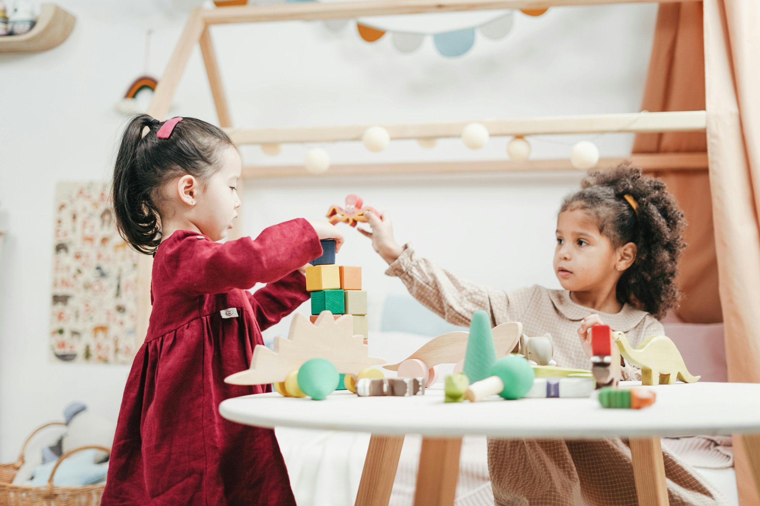 Two young girls playing with wooden building blocks and toys at a table in a playroom decorated with bunting and wall art.