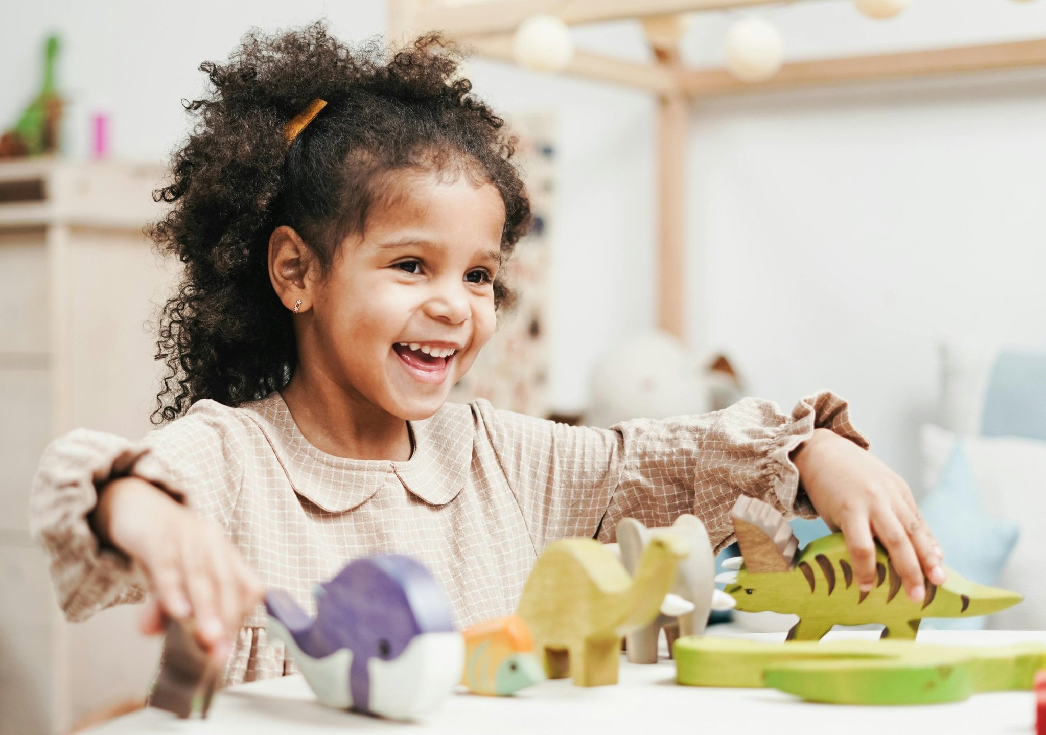 A young girl with curly hair and earrings happily playing with colorful wooden animal toys, including a purple zebra and a yellow tiger, on a white table in a bright room.