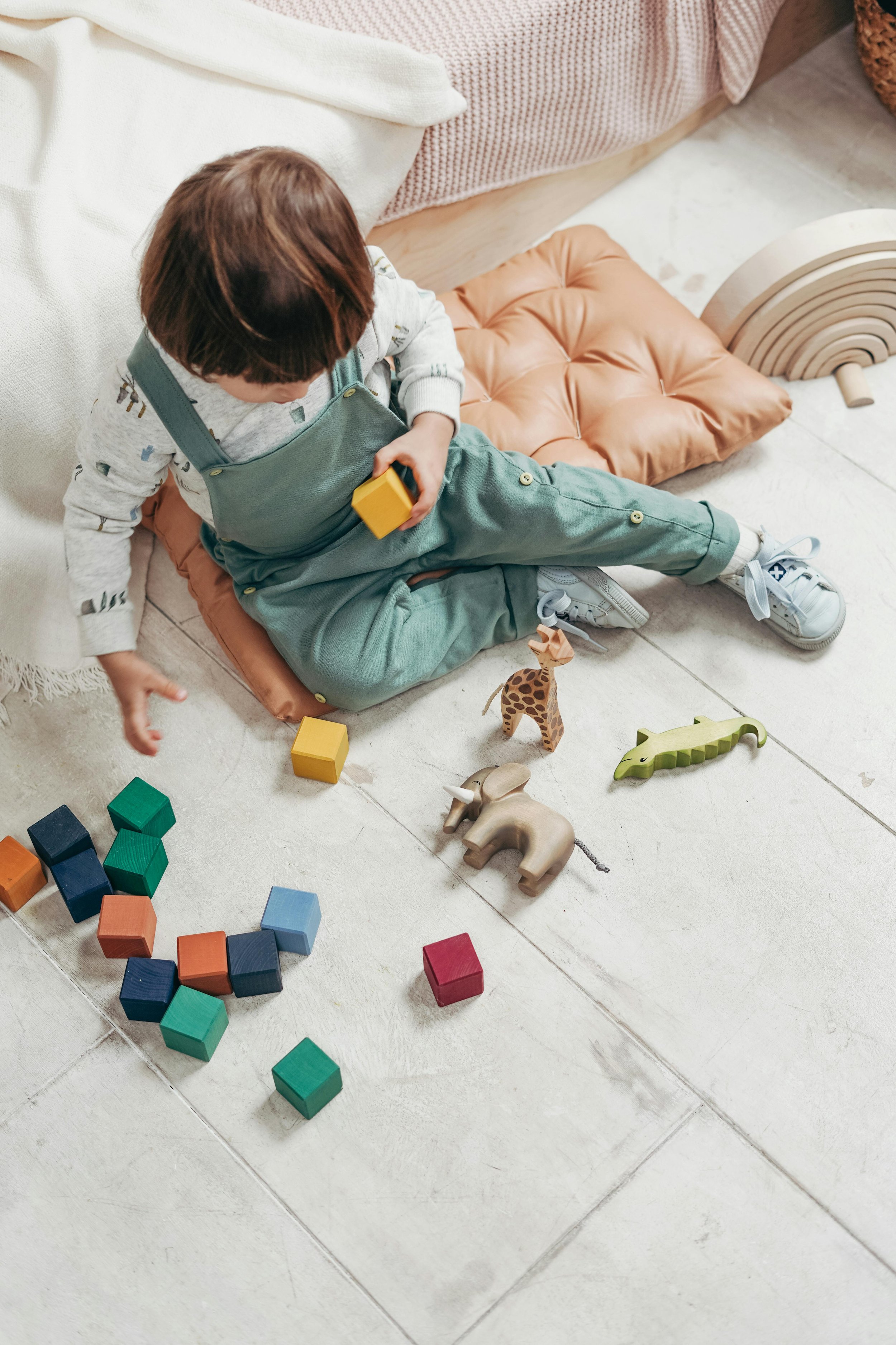 A young child sitting on a tan cushion on the floor, playing with colorful wooden blocks and animal toys, including an elephant, a giraffe, and a crocodile.