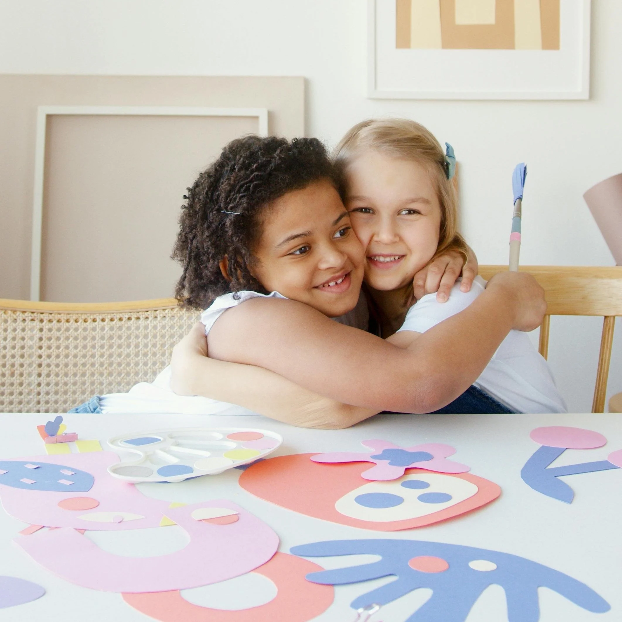 Two young girls hugging and smiling at camera, colorful paper cutouts on table, white wall with art in background.