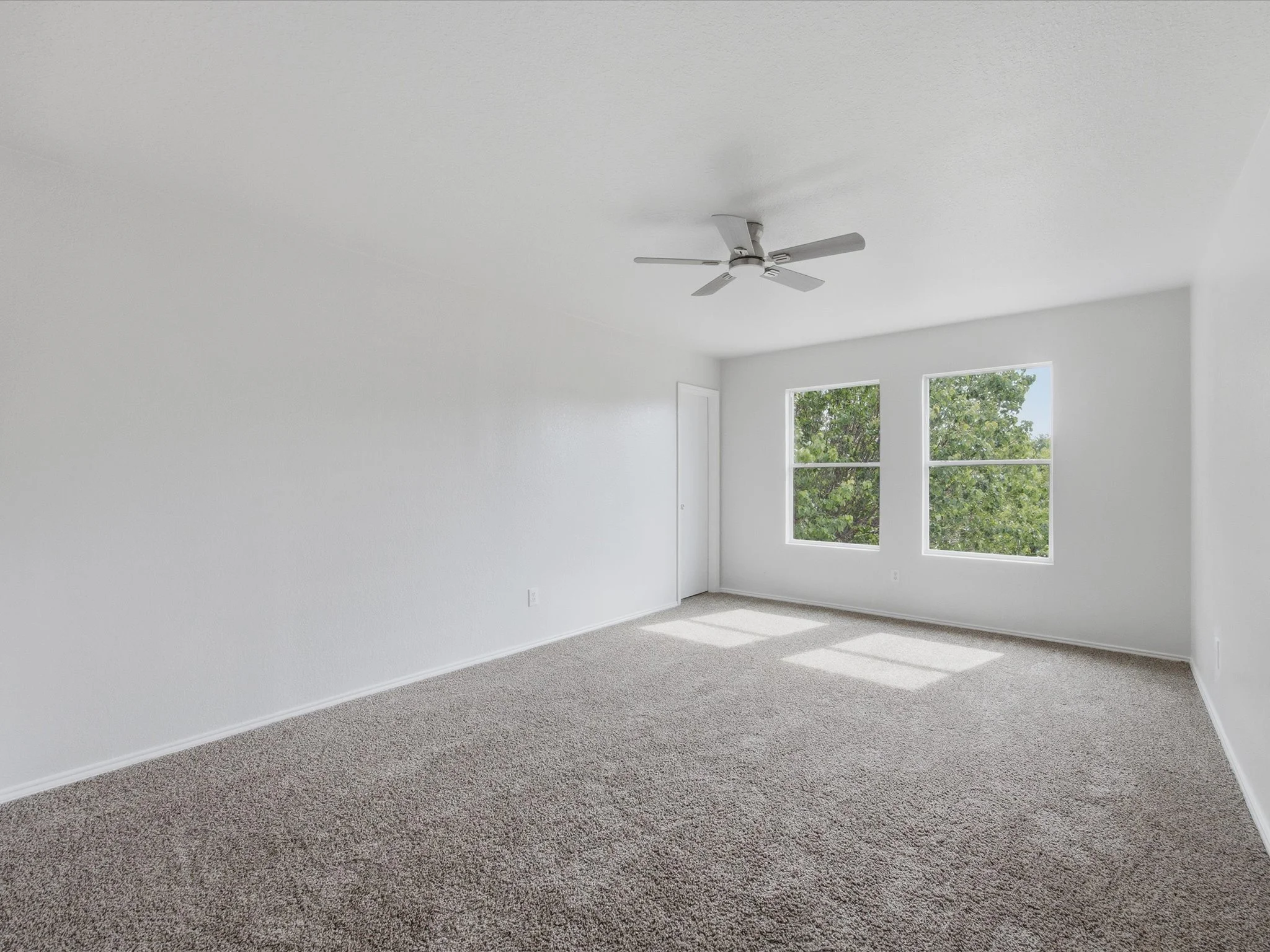 Empty room with white walls, gray carpet, two large windows showing green trees outside, and a ceiling fan.