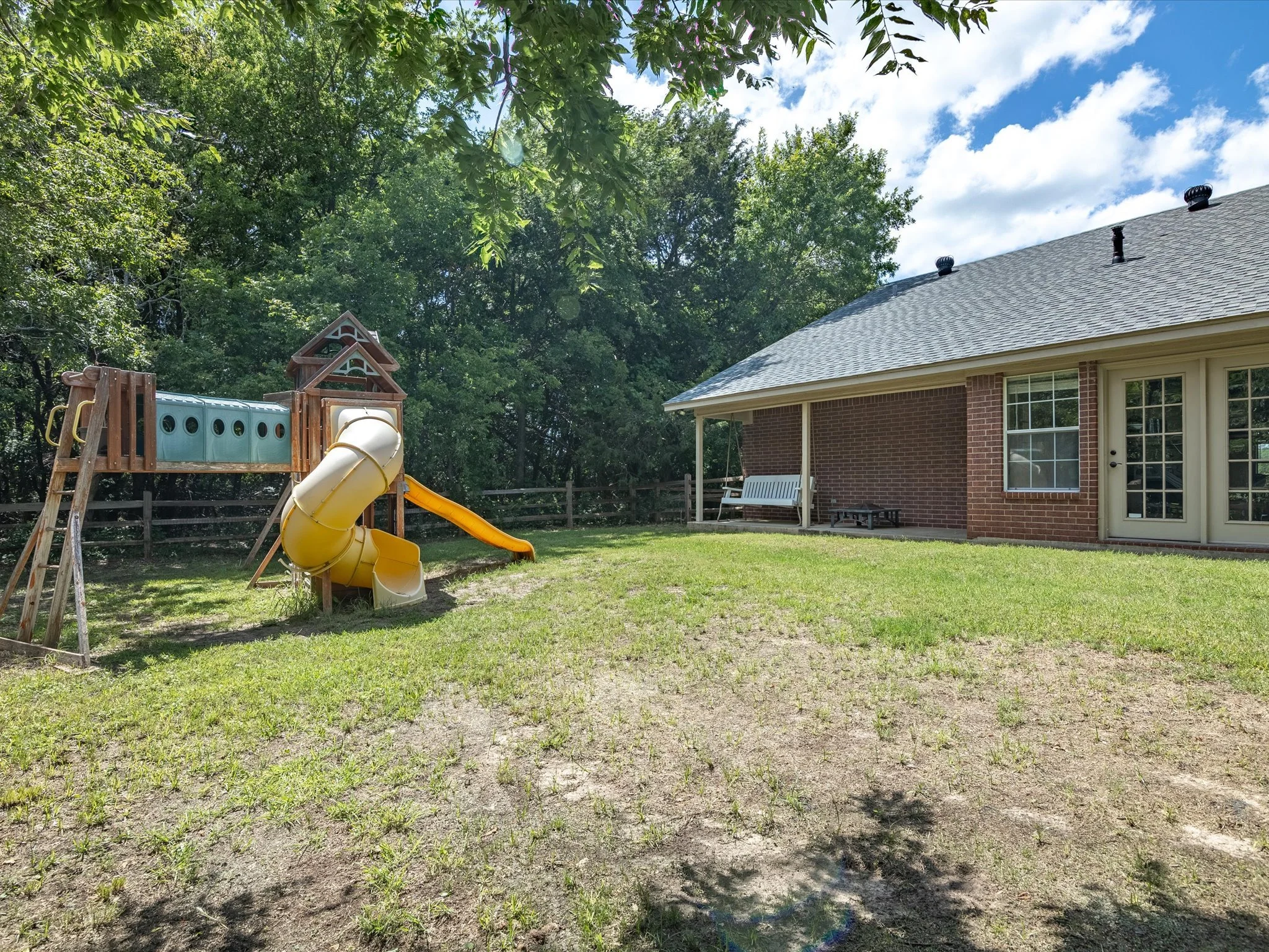 Backyard with a wooden playset, slide, and fenced yard. A house with brick walls, a gray shingled roof, and a screened porch is visible on the right. Large trees surround the yard, casting shadows on the grass.