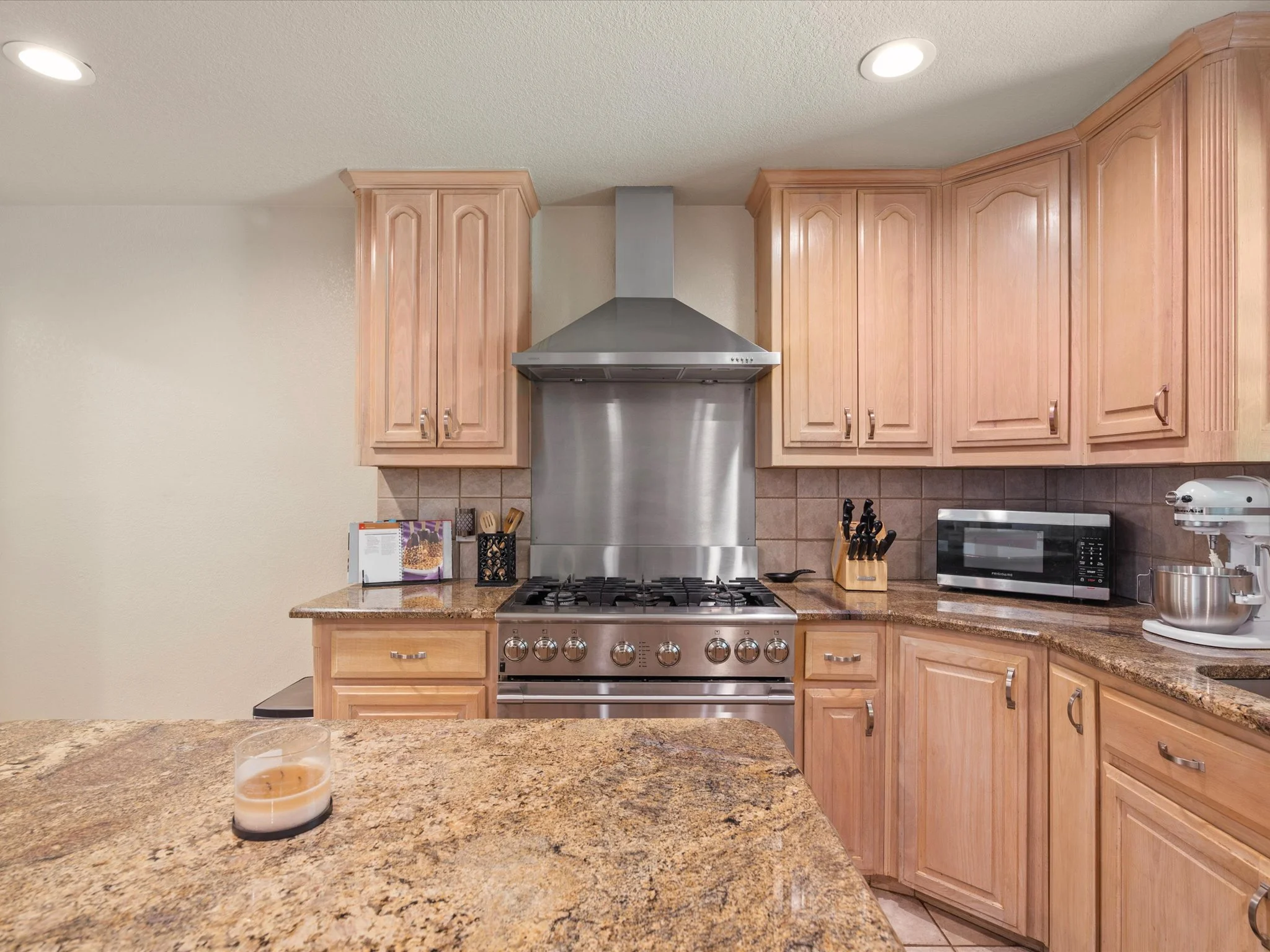 A kitchen with wooden cabinets, a stainless steel range hood above a stove, a microwave, and a countertop with a candle, knives, and kitchen appliances.