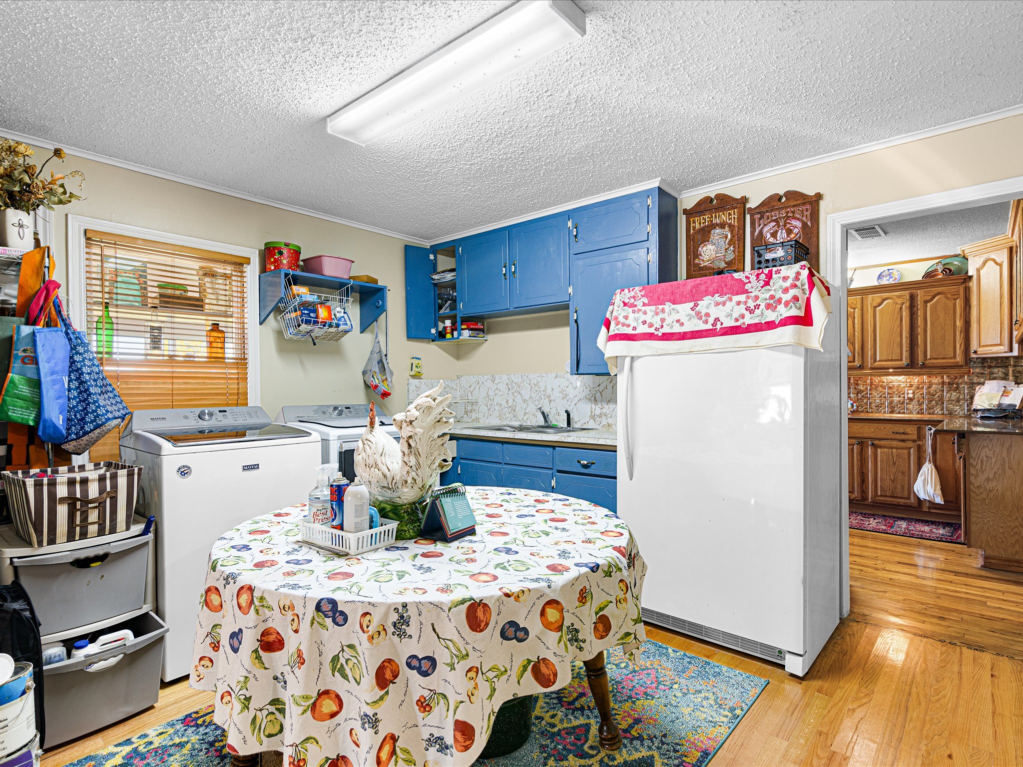 A kitchen with blue cabinets, a round table covered with a fruit-themed tablecloth, and various kitchen items, including a washing machine, a dish drying rack, and wooden cabinets.