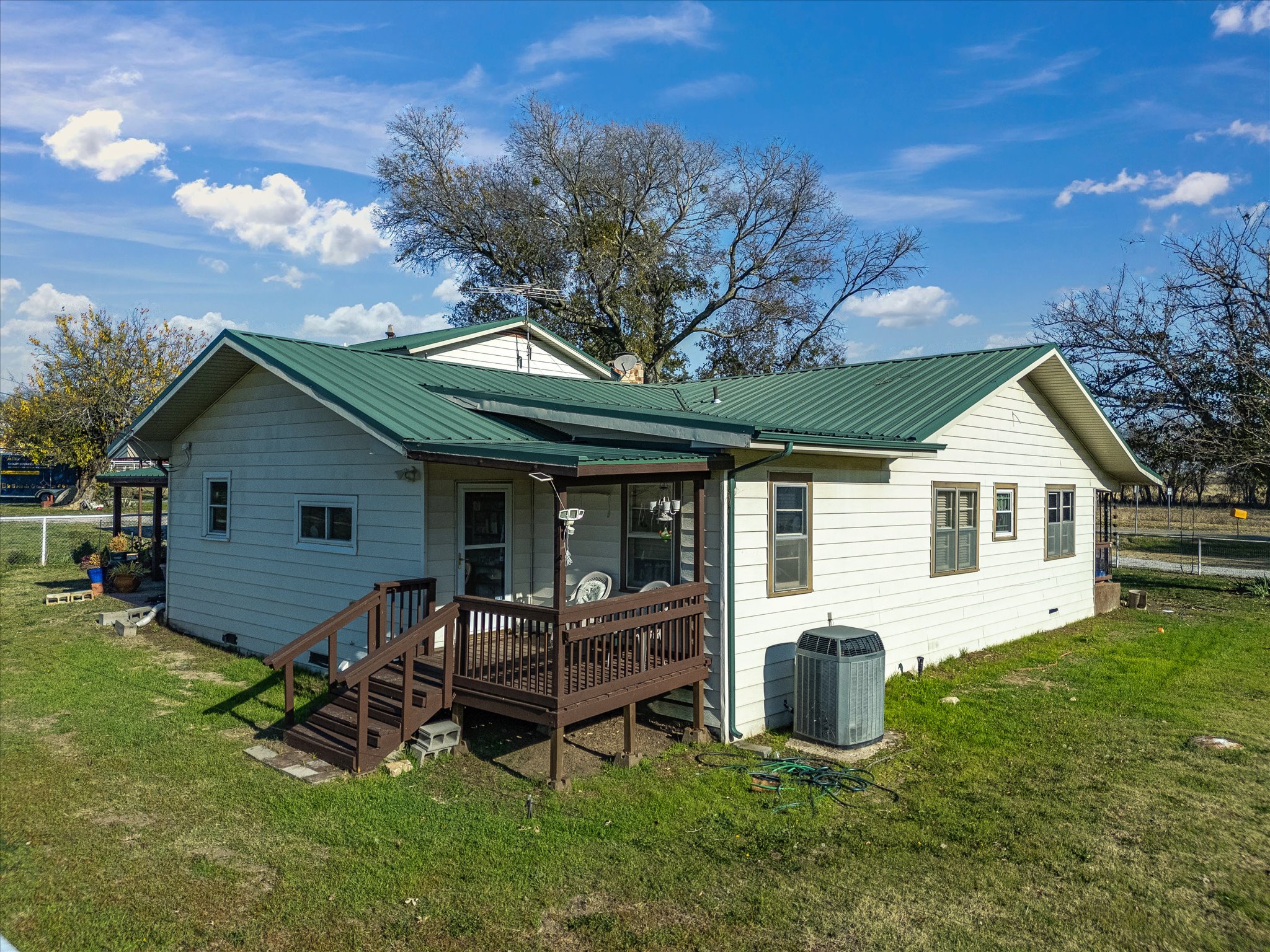 A two-story house with a green metal roof, white siding, and a small wooden porch with stairs. There are trees without leaves behind and around the house, and a backyard with green grass. A fence and some equipment are visible in the background under