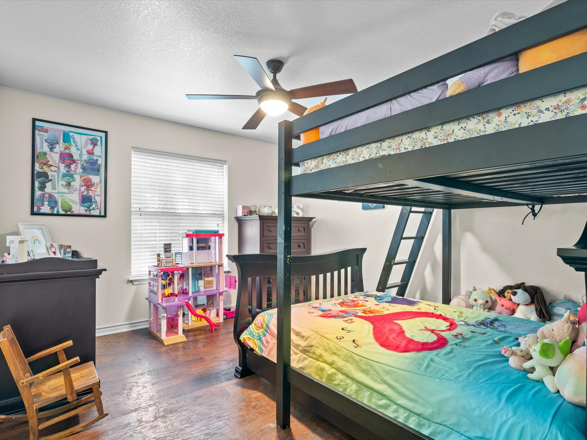 A bedroom featuring a bunk bed with a bedspread decorated with flamingos, plush toys on the bed, a chest of drawers, a framed poster of trolls on the wall, a window with blinds, and a daybed with a pillow and quilt.
