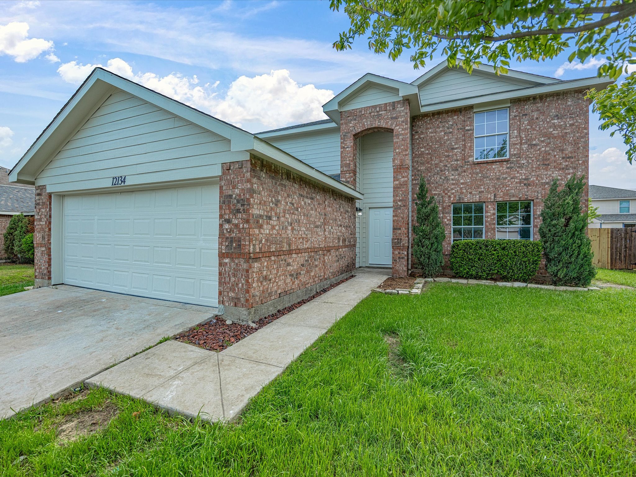 Brick and light siding two-story house with attached garage, concrete walkway, front lawn with bushes, and trees under blue sky with clouds.