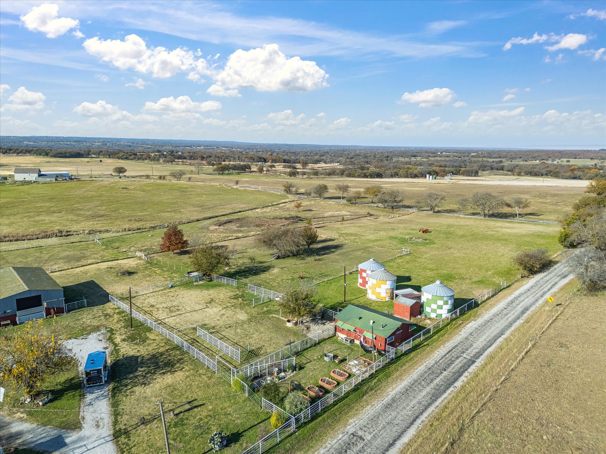 A rural farm scene with green pastures, trees, and farm buildings. A red barn and colorful silo structures are in the foreground, with open fields and a cloudy blue sky in the background.