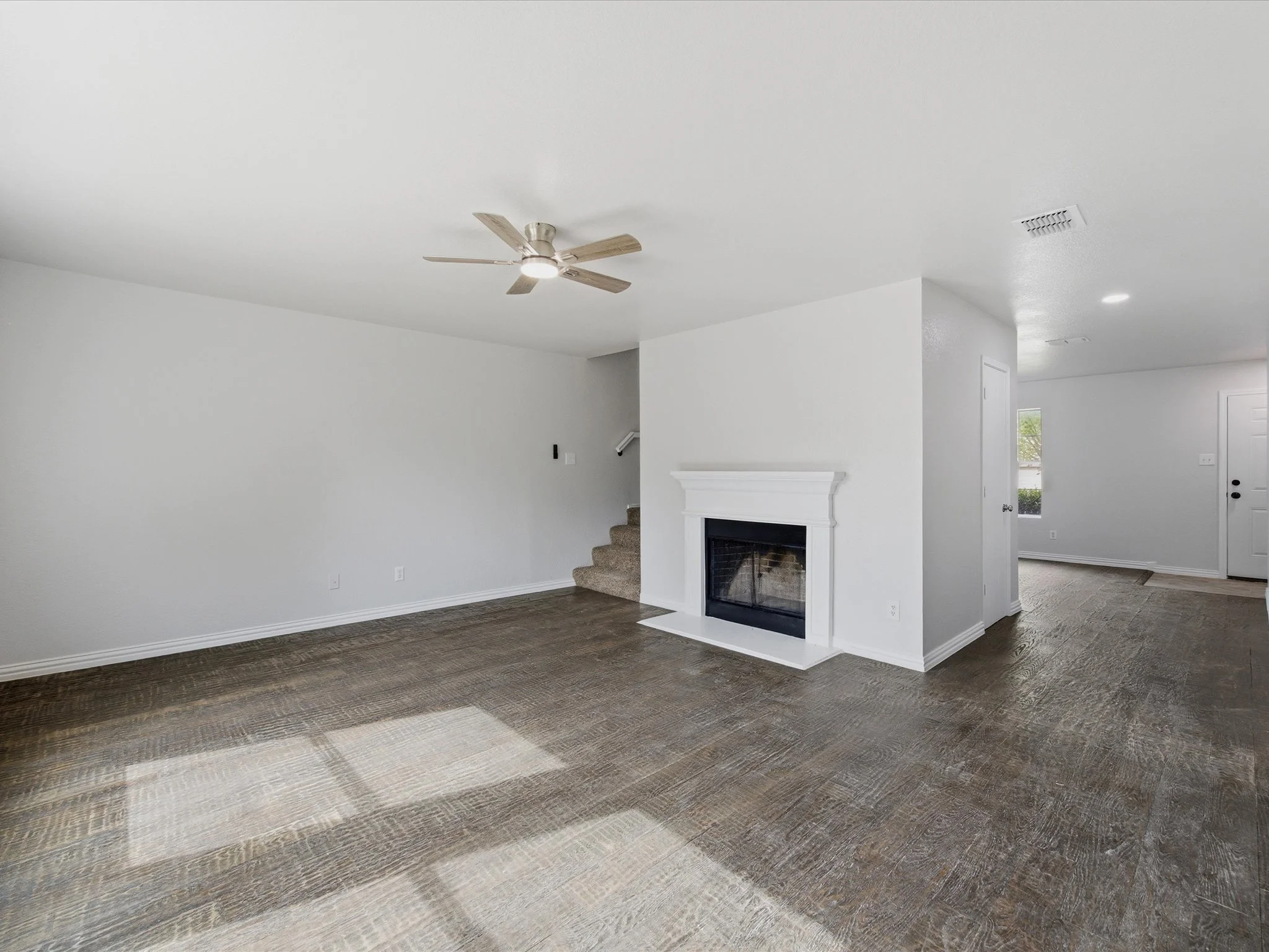 Empty living room with white walls, dark brown hardwood flooring, a white fireplace, ceiling fan with five wooden blades, and natural light coming through a window.