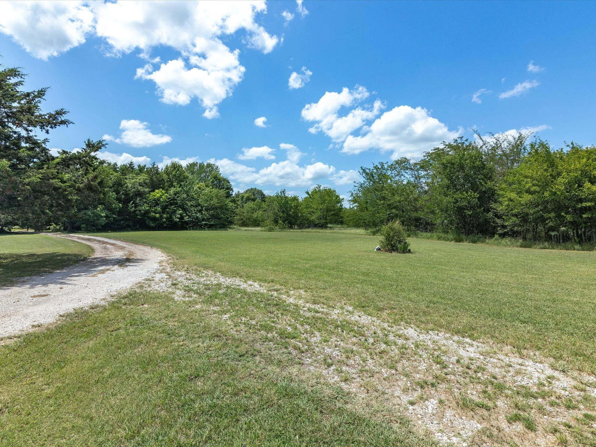 A gravel path through a grassy field surrounded by trees under a blue sky with scattered white clouds.