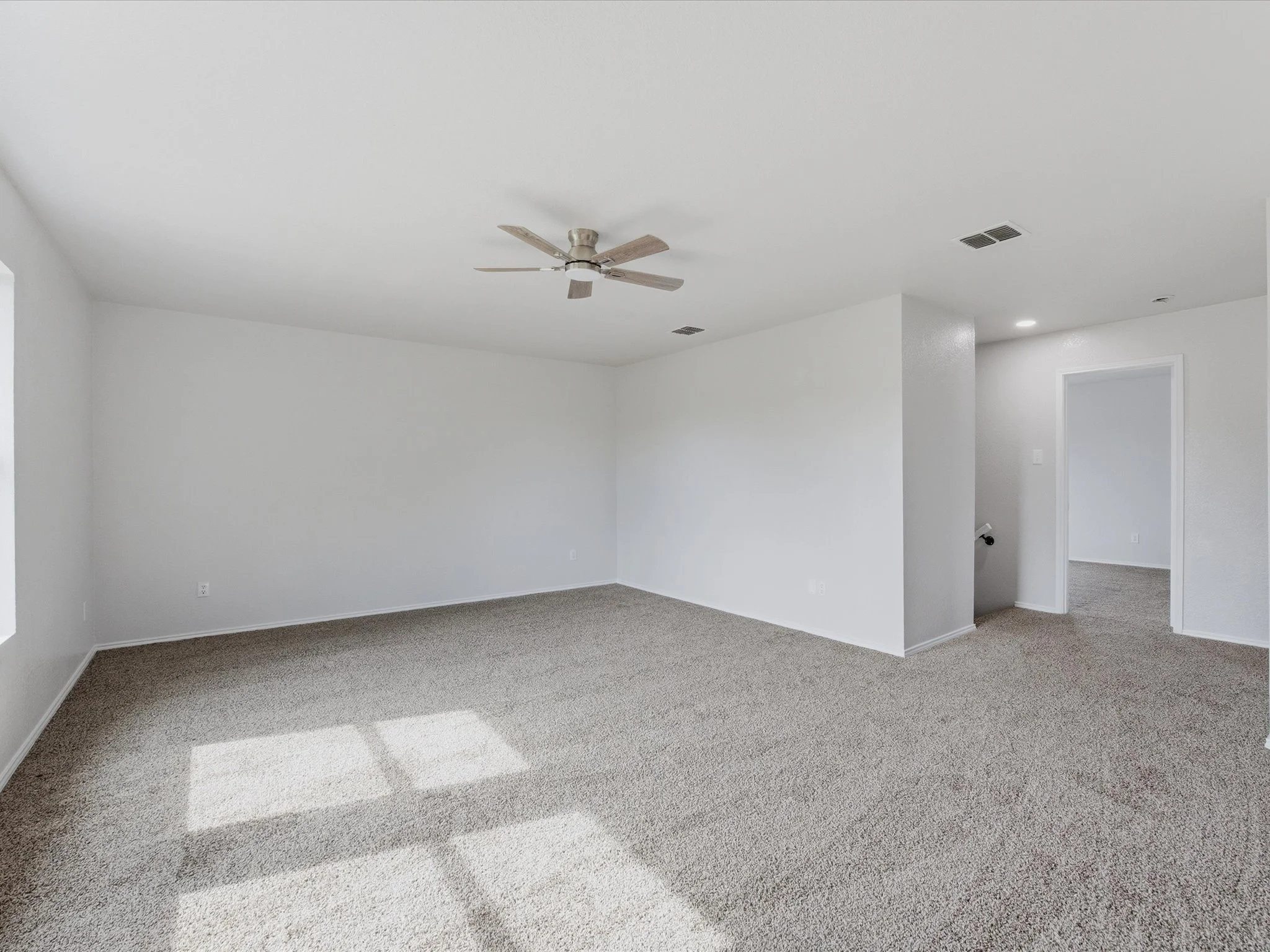 Empty living room with beige carpet, white walls, ceiling fan, and a doorway in the corner.