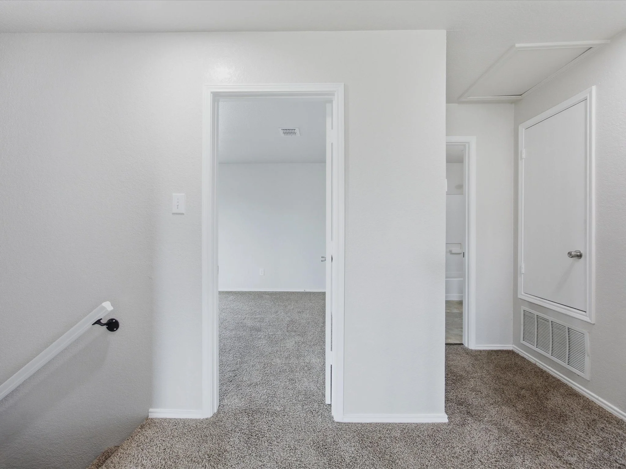 View of an empty interior hallway in a residential home with introduction of doorways, textured light gray walls, beige carpeted floors, and a ceiling vent.