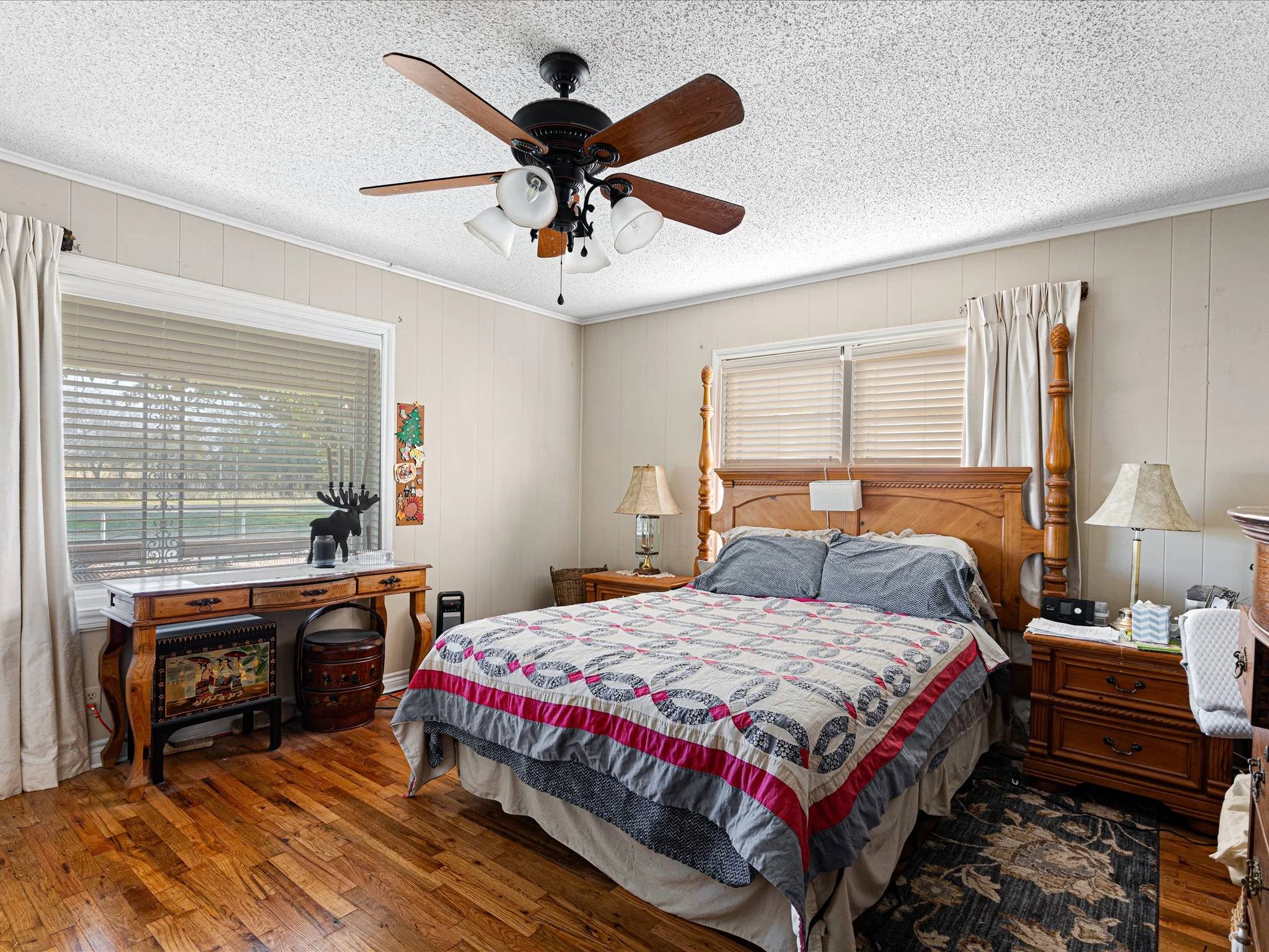 A bedroom with a wooden bed, a quilted bedspread, and two lamps. The room has three windows with blinds and curtains, and a ceiling fan with lights.