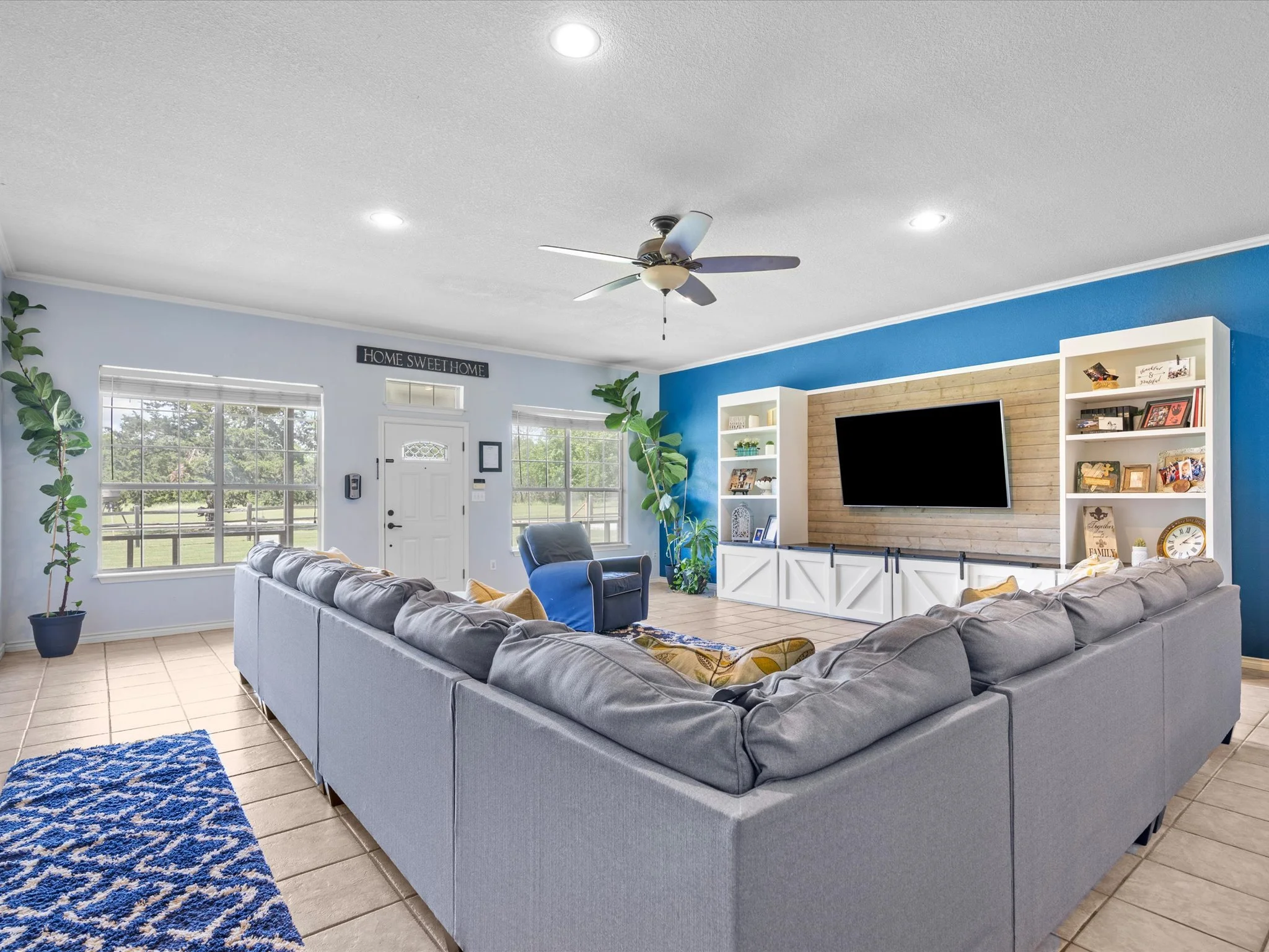 A living room with a gray sectional sofa, a blue armchair, a wooden floor, a blue and white rug, large windows, a ceiling fan, a wall-mounted TV, white shelves with decorations, and a door with a small window.