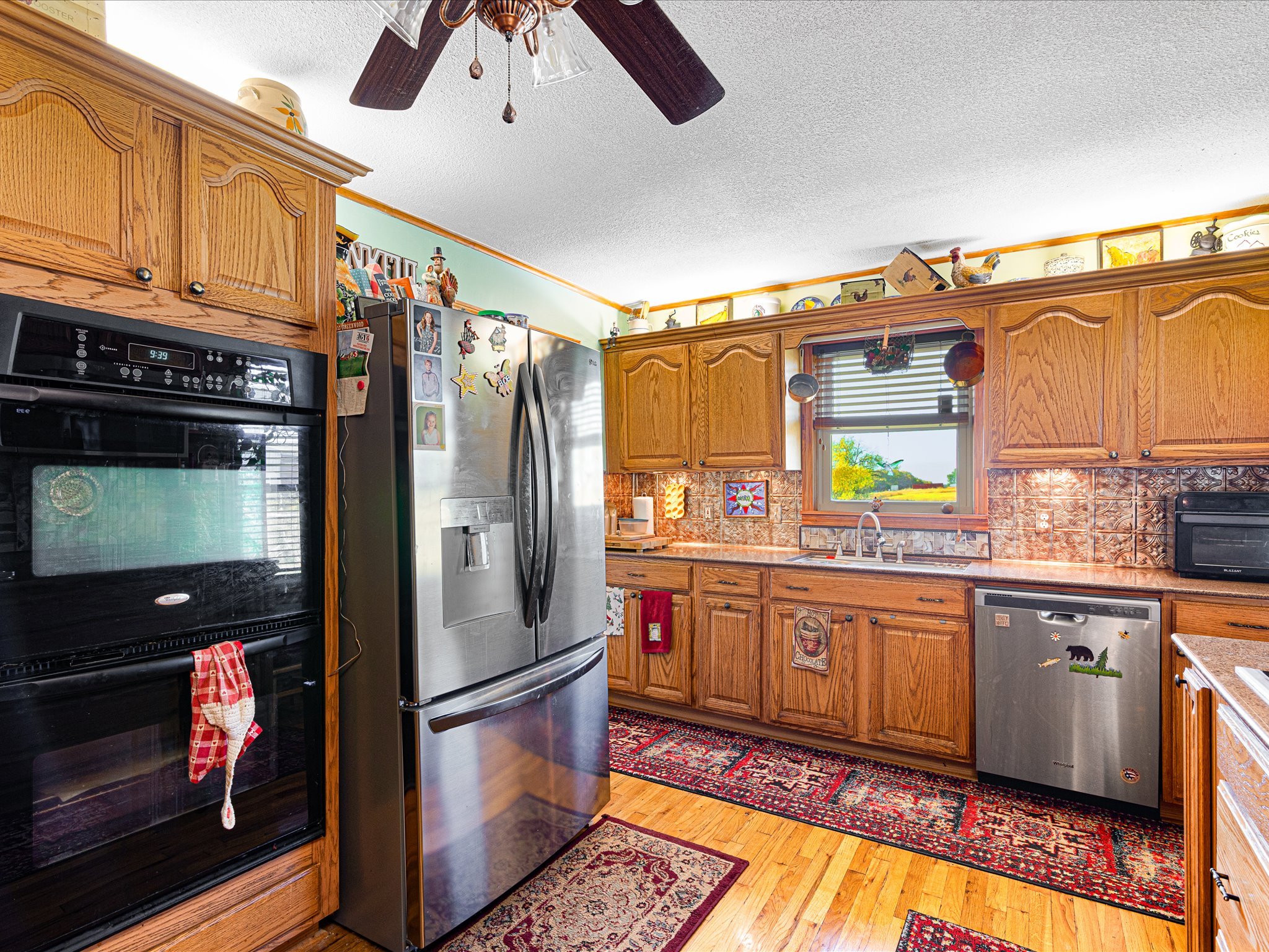 A kitchen with wooden cabinets, a black oven, a stainless steel refrigerator with photos and magnets, a window with blinds, and a colorful area rug on a wooden floor.