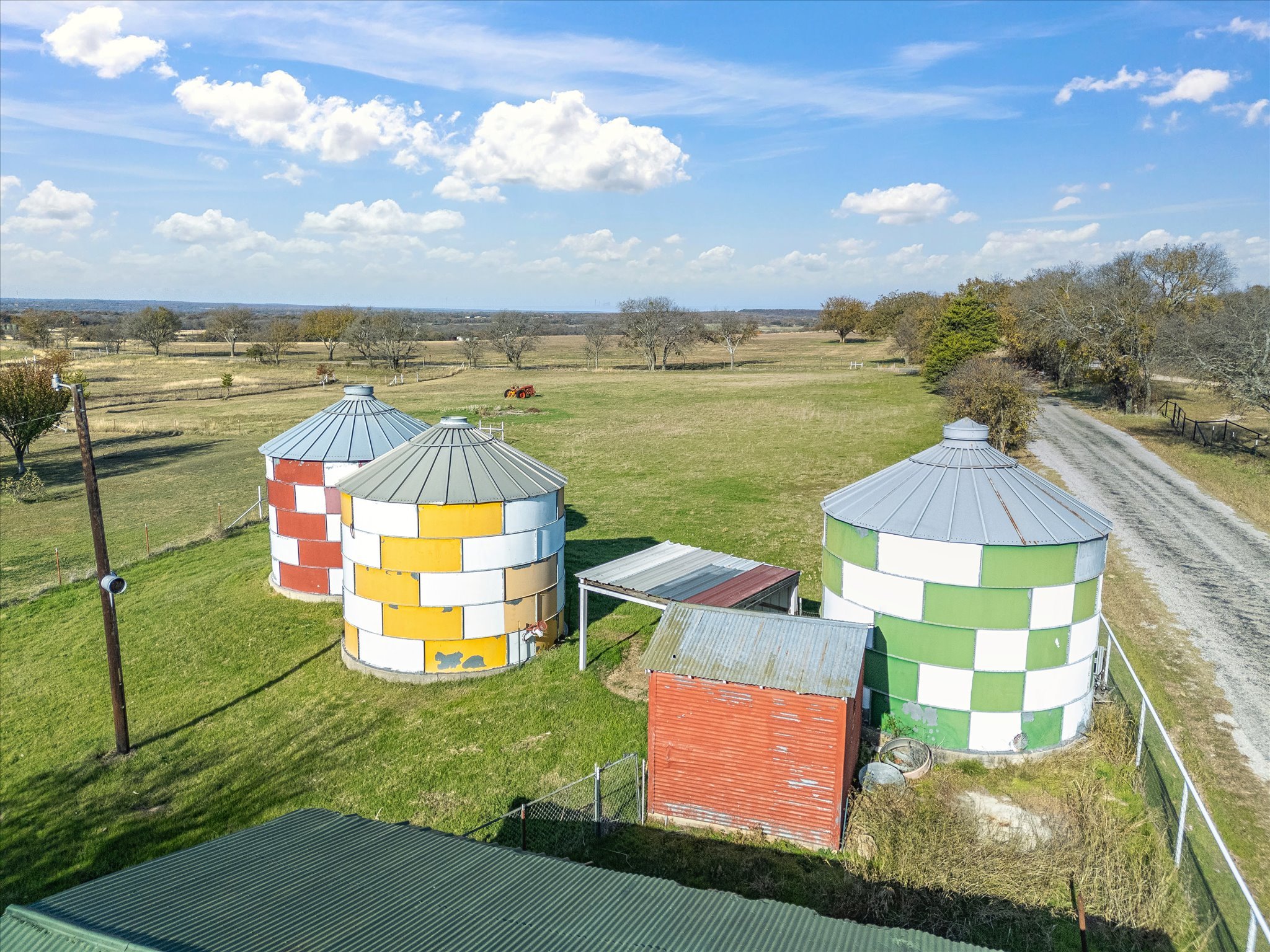 Colorful farm silos with a red building at the bottom, green grass, a dirt pathway, trees, and a blue sky with scattered clouds.