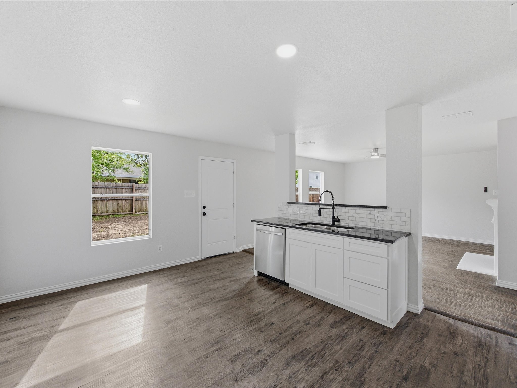 Interior view of a bright, empty kitchen with white walls, a white kitchen island with black countertop, and wood flooring. There are windows showing a backyard with a wooden fence and green trees.