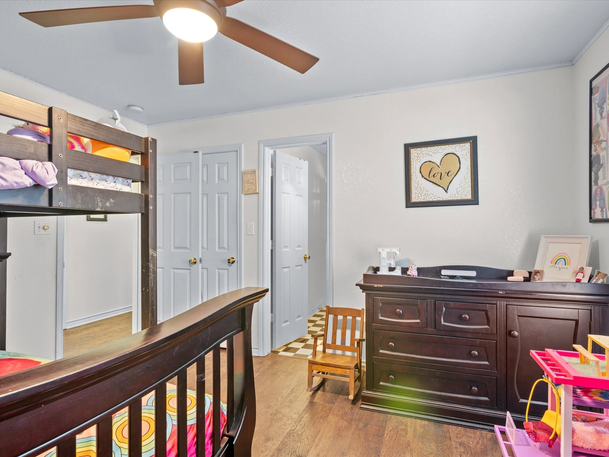 A nursery with a black wooden bunk bed, framed artwork on cream walls, a wooden dresser, and a small wooden chair.