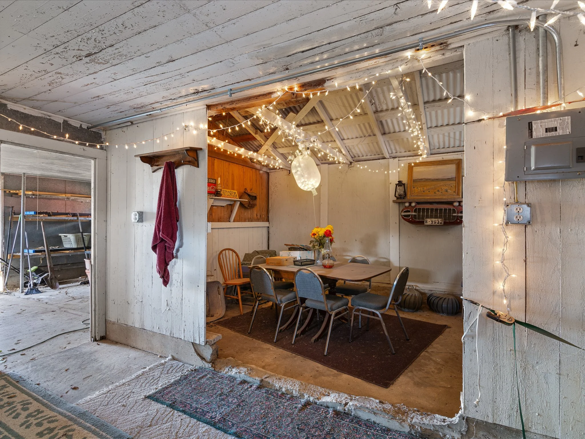 A rustic dining area with a long wooden table and chairs, string lights overhead, and vintage decor on the walls, set inside a partially exposed barn or workshop with weathered walls and ceiling.