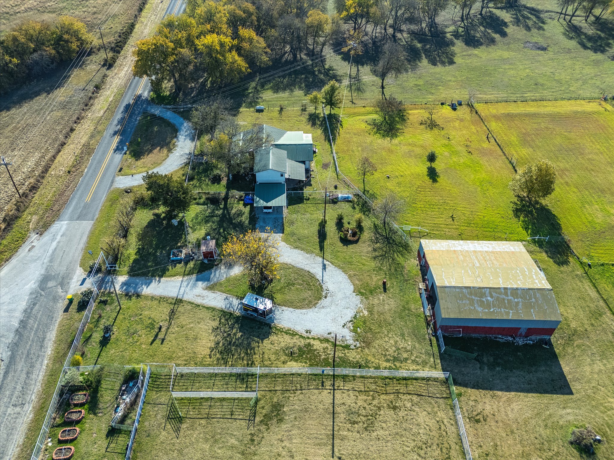 An aerial view of a rural property with a house, trees, fenced yards, a driveway, and surrounding open fields. A road runs alongside the property with power lines overhead.