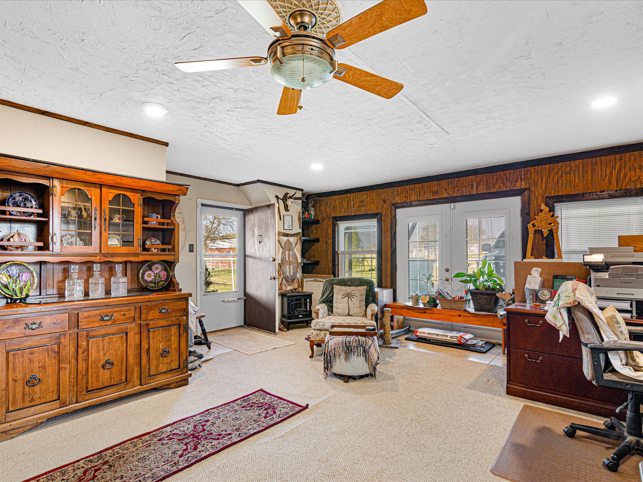 Living room with a ceiling fan, wooden furniture, a chair, a small table, and various decorative items, with sunlight coming through multiple windows.