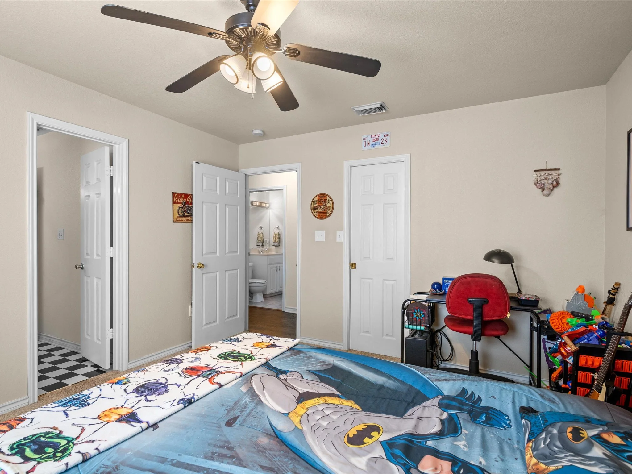 A room with beige walls and white doors, decorated with children's posters and shelved toys. The ceiling has a black fan with lights. An open door leads to a bathroom. On the floor, there are Batman-themed bed sheets, a red chair, a floor lamp, and s