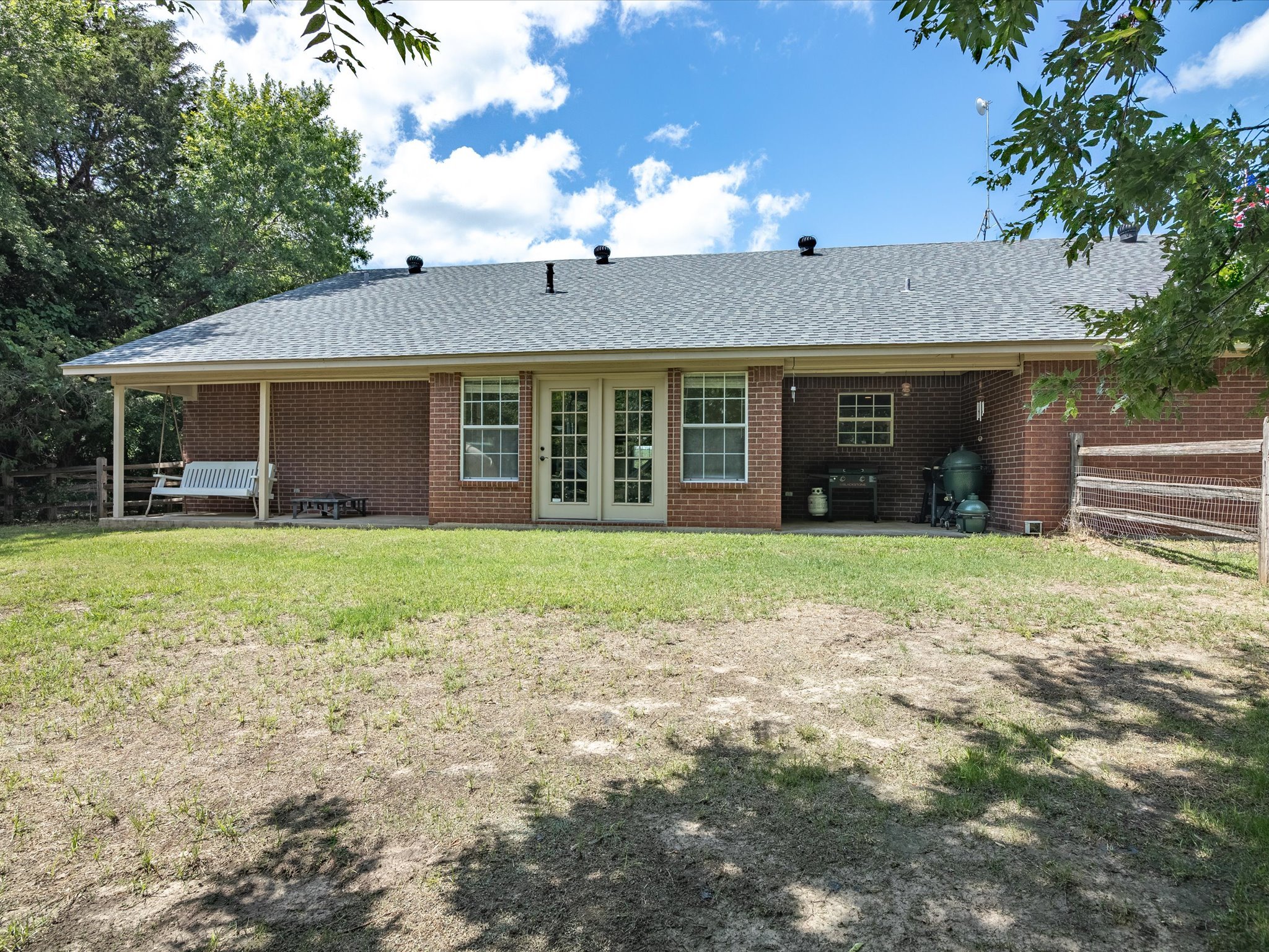 Backyard view of a brick house with a gray shingle roof, white double doors, two windows, a porch with a swing, a grill, and a fenced area. Surrounding trees and a partly cloudy sky.