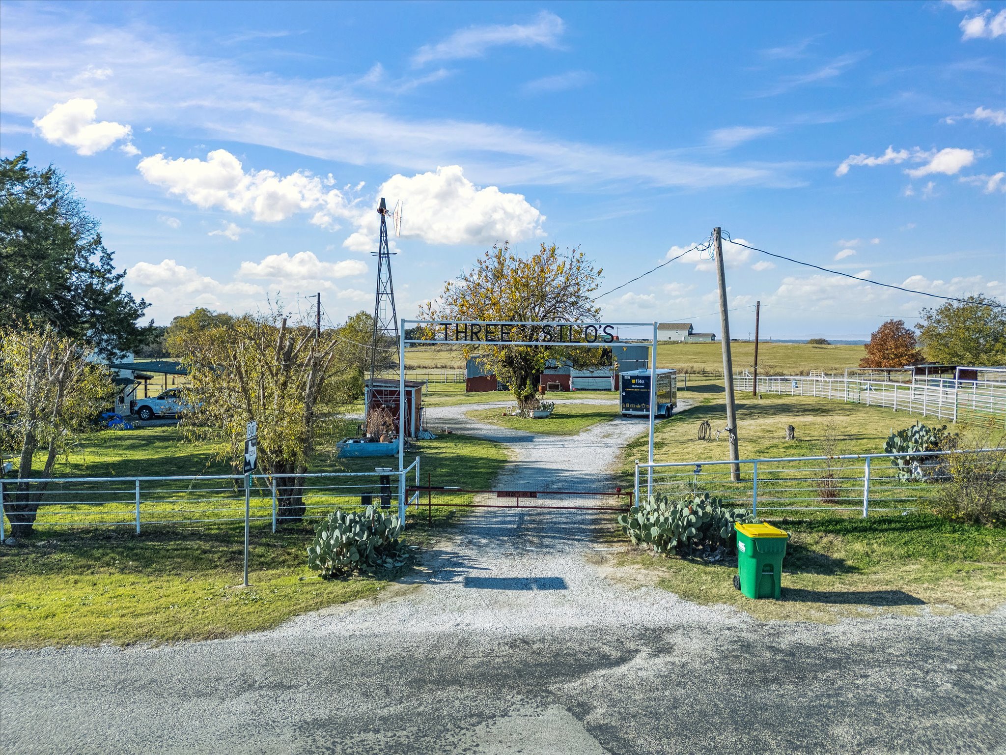 A rural landscape with a gravel driveway, a fenced yard with trees and cactus, a sign for 'Threelog's', and a windmill in the background against a blue sky with clouds.