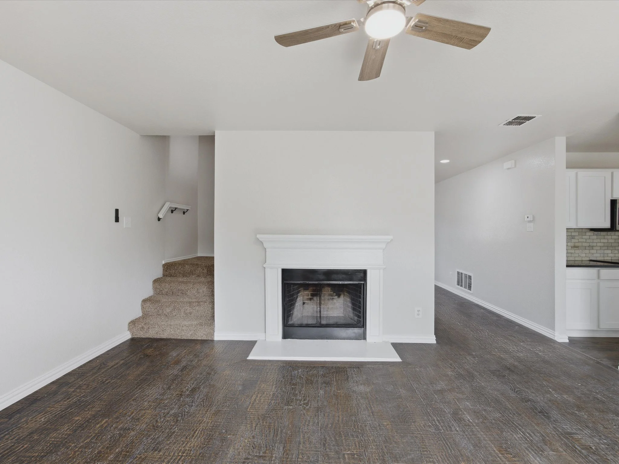Empty living room with white walls, a fireplace, ceiling fan, dark hardwood floors, and a partial view of a kitchen.