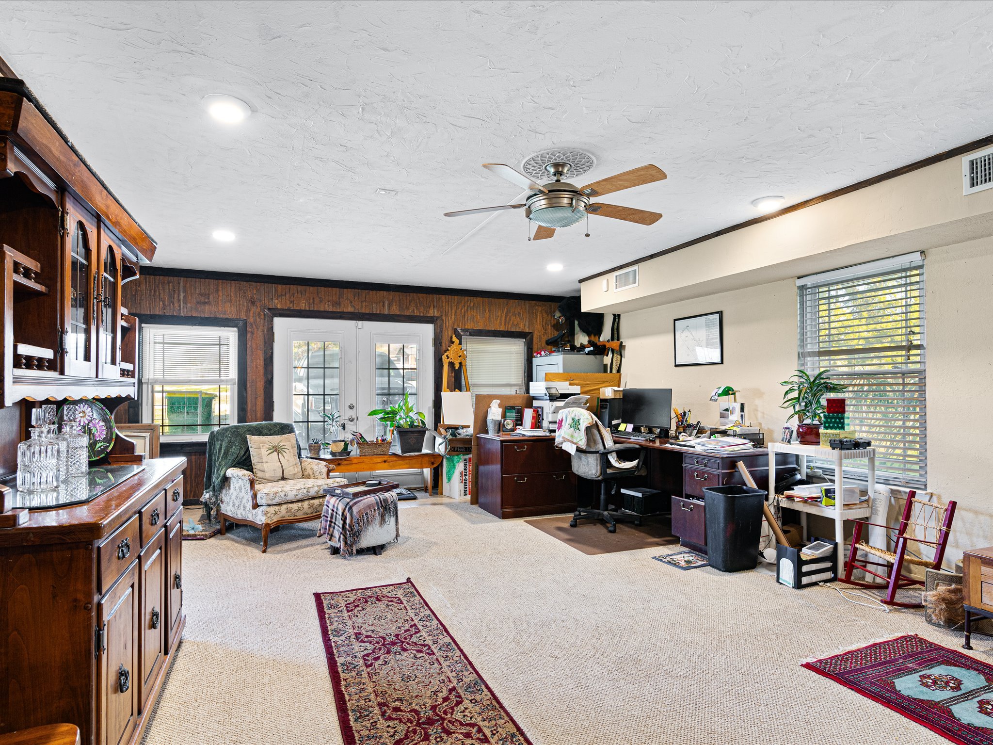 A cluttered home office with various furniture, computers, and decorations, large window with blinds, ceiling fan, and wooden accents.