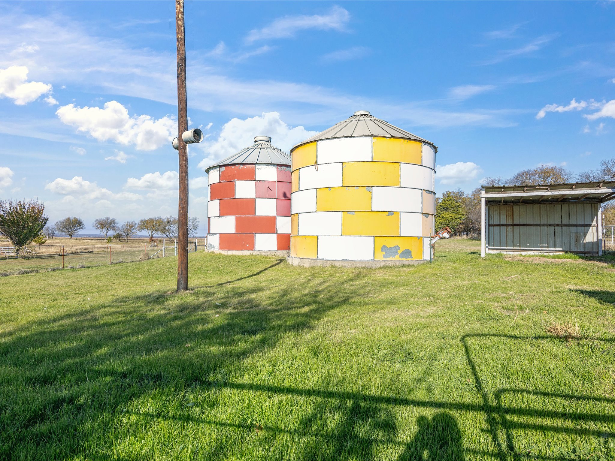 Two cylindrical grain silos with metal roofs, one painted red and white checkered and the other yellow and white checkered, standing on a grassy field under a blue sky with scattered clouds. A utility pole with a light or camera attached is in the fo