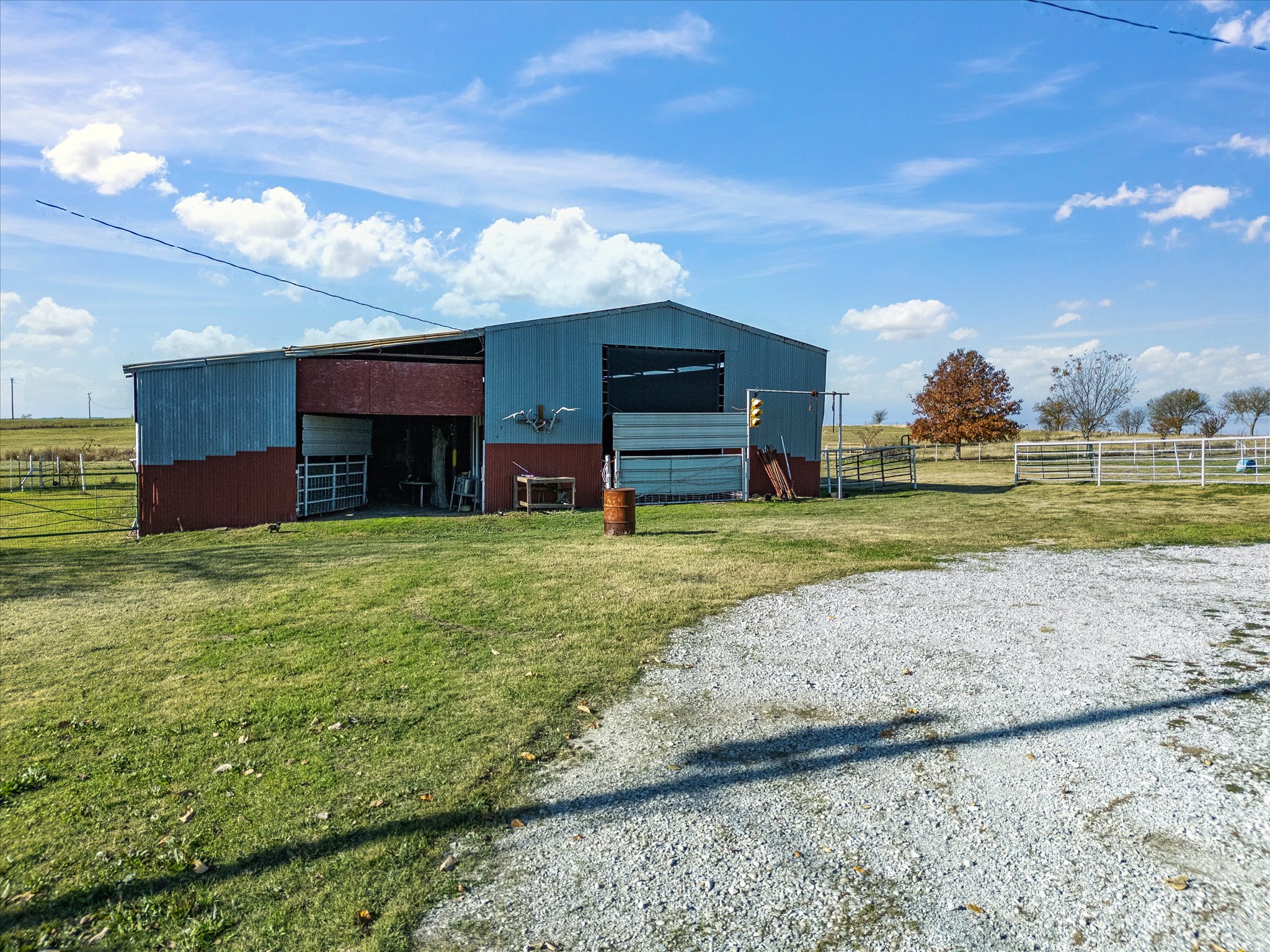 A farm building with blue and red ribbed metal siding, open on one side, with a green field and trees in the background, under a partly cloudy blue sky.