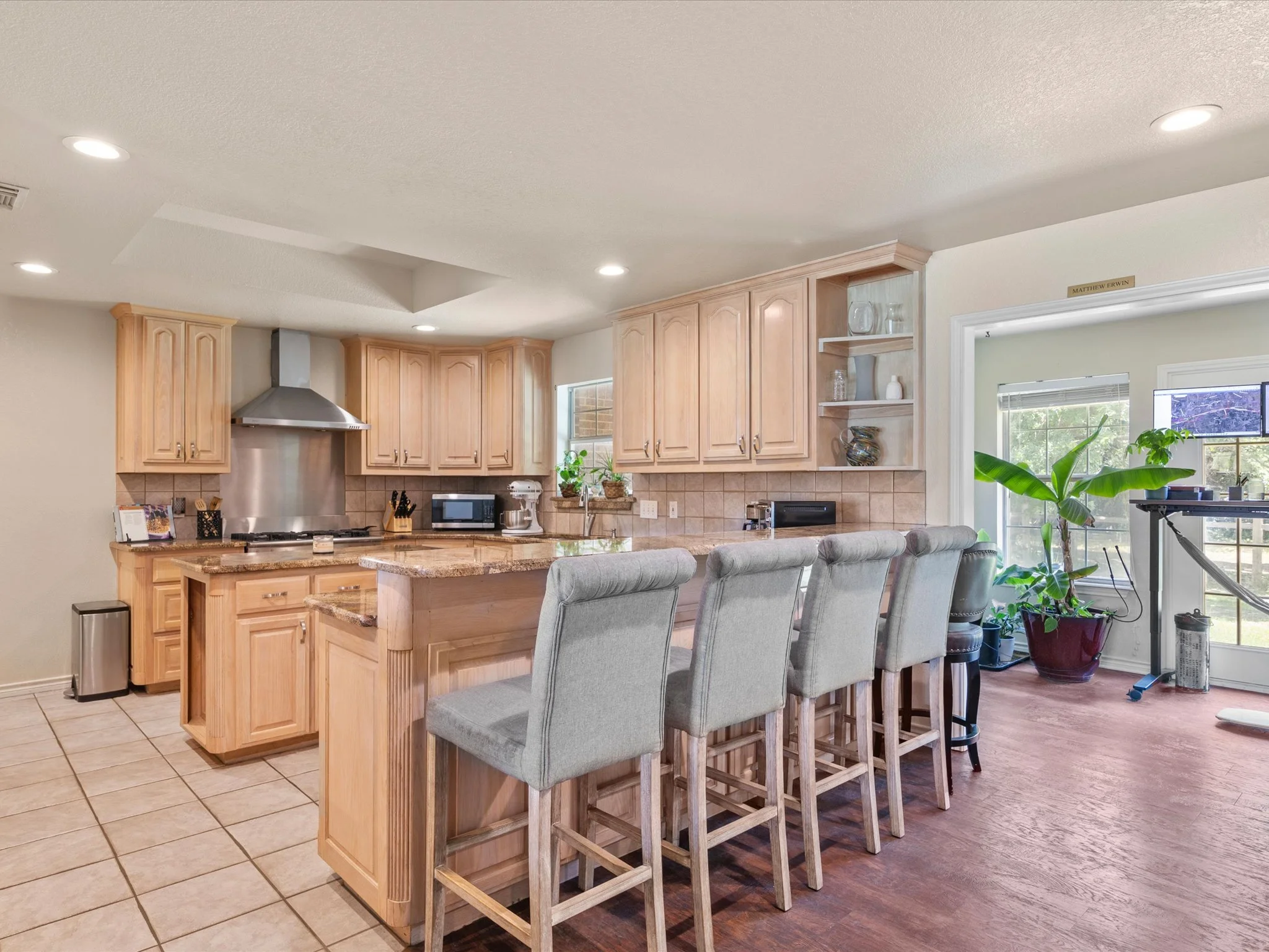 Interior view of a kitchen with light wood cabinets, a countertop with kitchen appliances, and a dining area with gray upholstered chairs at a wooden counter with a bar top. There are plants near a large window and a sliding door.