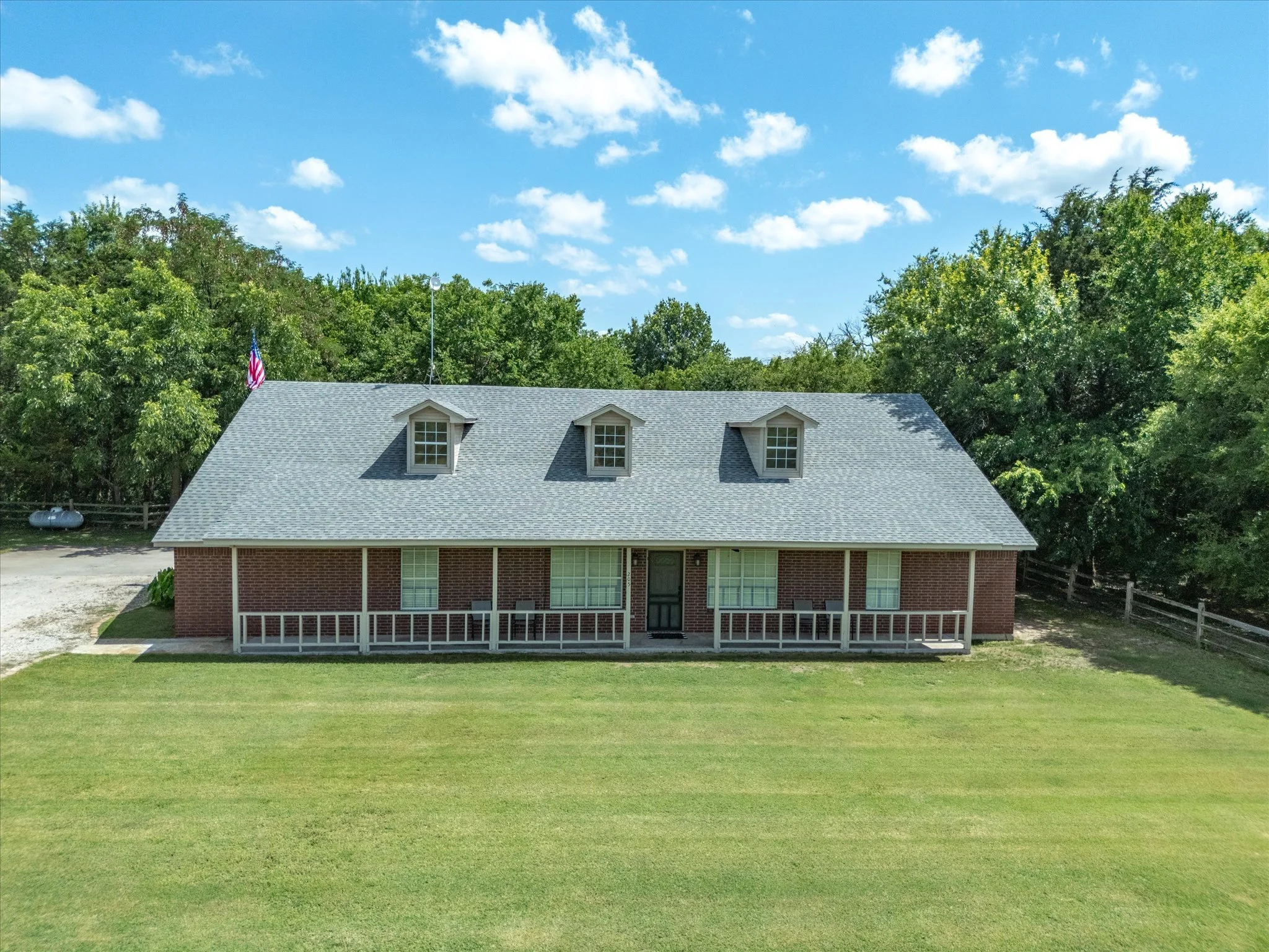 View of a house with a gray shingled roof, three dormer windows, a brick exterior, a small porch, and a large well-maintained lawn in the foreground. The house is surrounded by tall trees and under a bright blue sky with scattered white clouds.